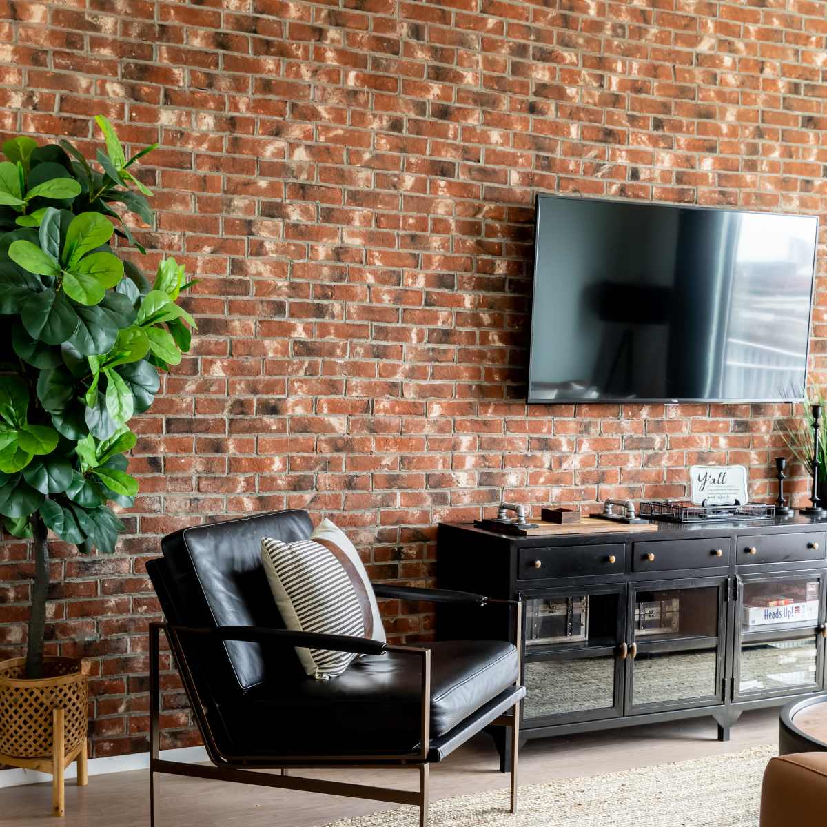 A modern living room with a black armchair, a television mounted on a brick wall, and a potted plant beside a dark wooden console table.