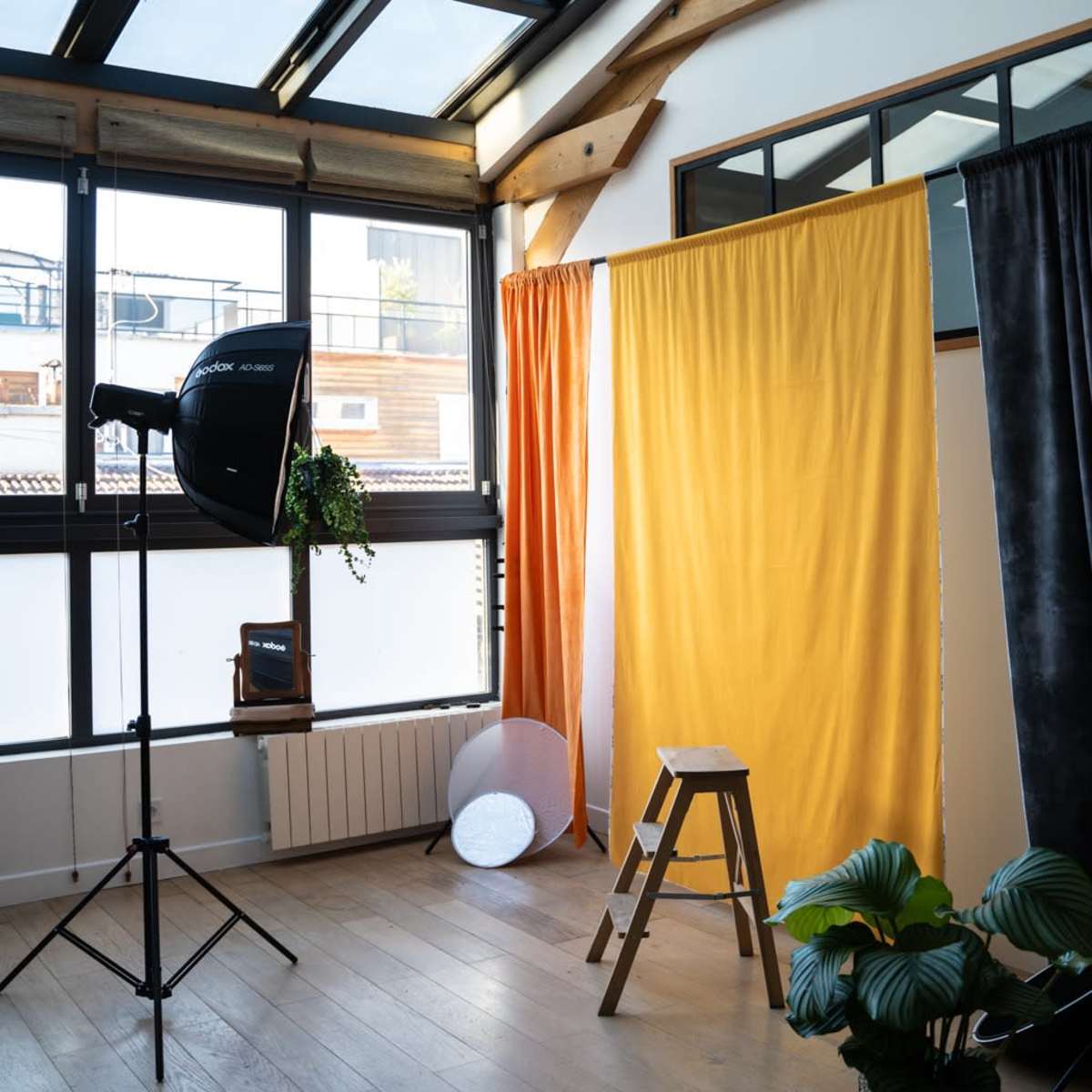 A well-lit studio space with a wooden floor, a photography backdrop with orange and yellow curtains, a stool, a light stand, and a potted plant.