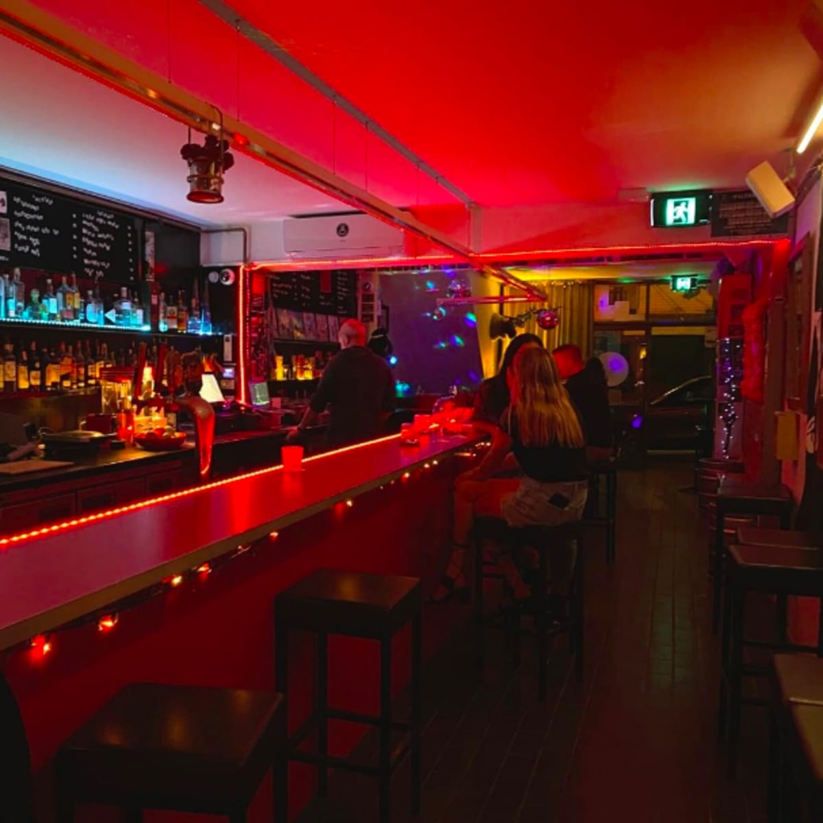 A dimly lit bar with red and blue lighting, featuring a long bar counter and patrons sitting on stools.
