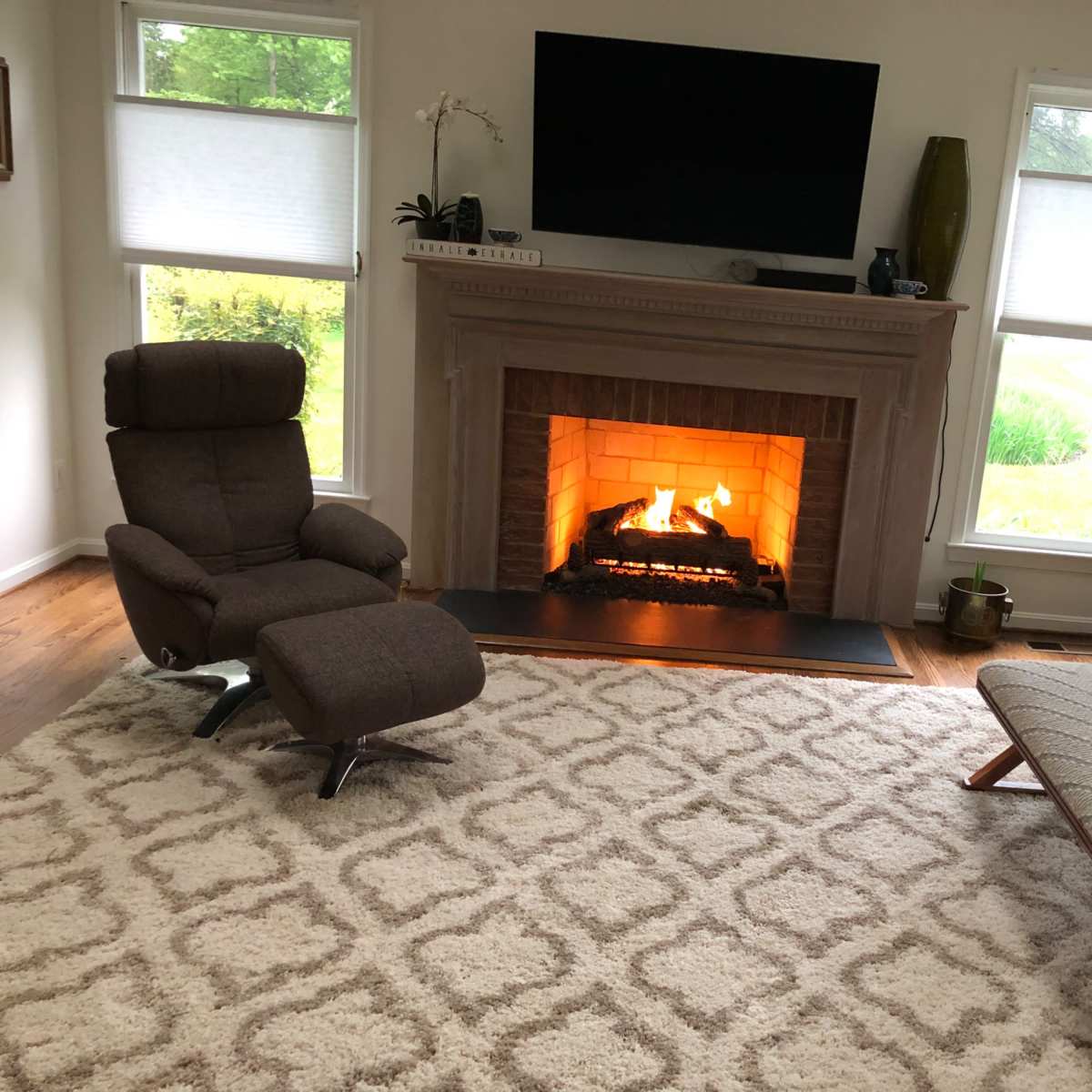 A cozy living room with a fireplace, two chairs, a bench, a large rug, and a television mounted on the wall.