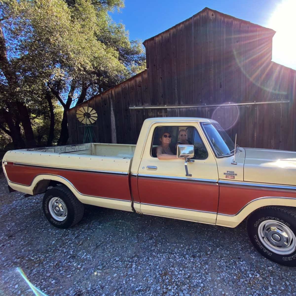 A vintage truck with a white and orange exterior is parked in front of a weathered barn under bright sunlight, with two people sitting inside.