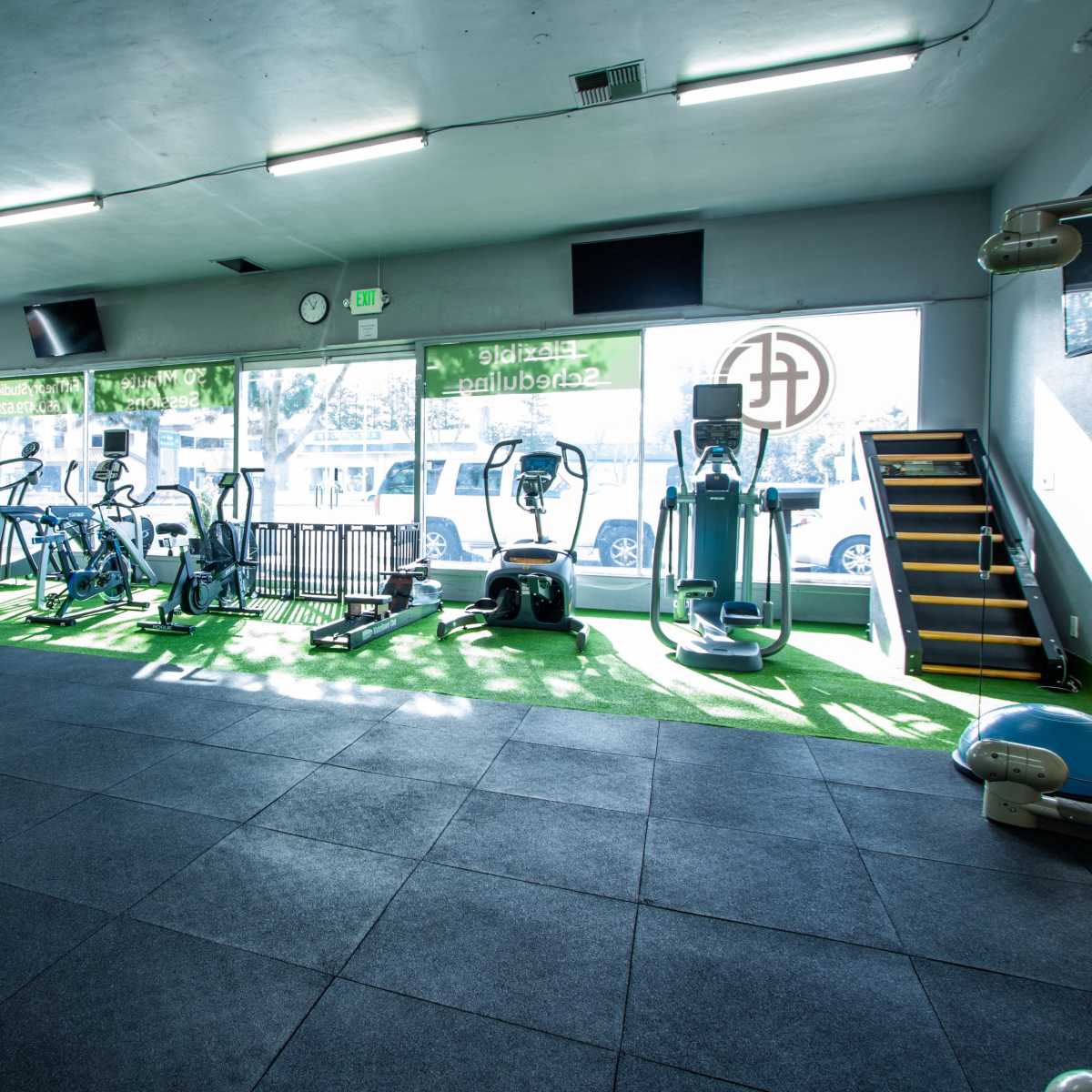 A gym interior with various exercise equipment, including cardio machines and weights, situated on rubber flooring and illuminated by natural light.
