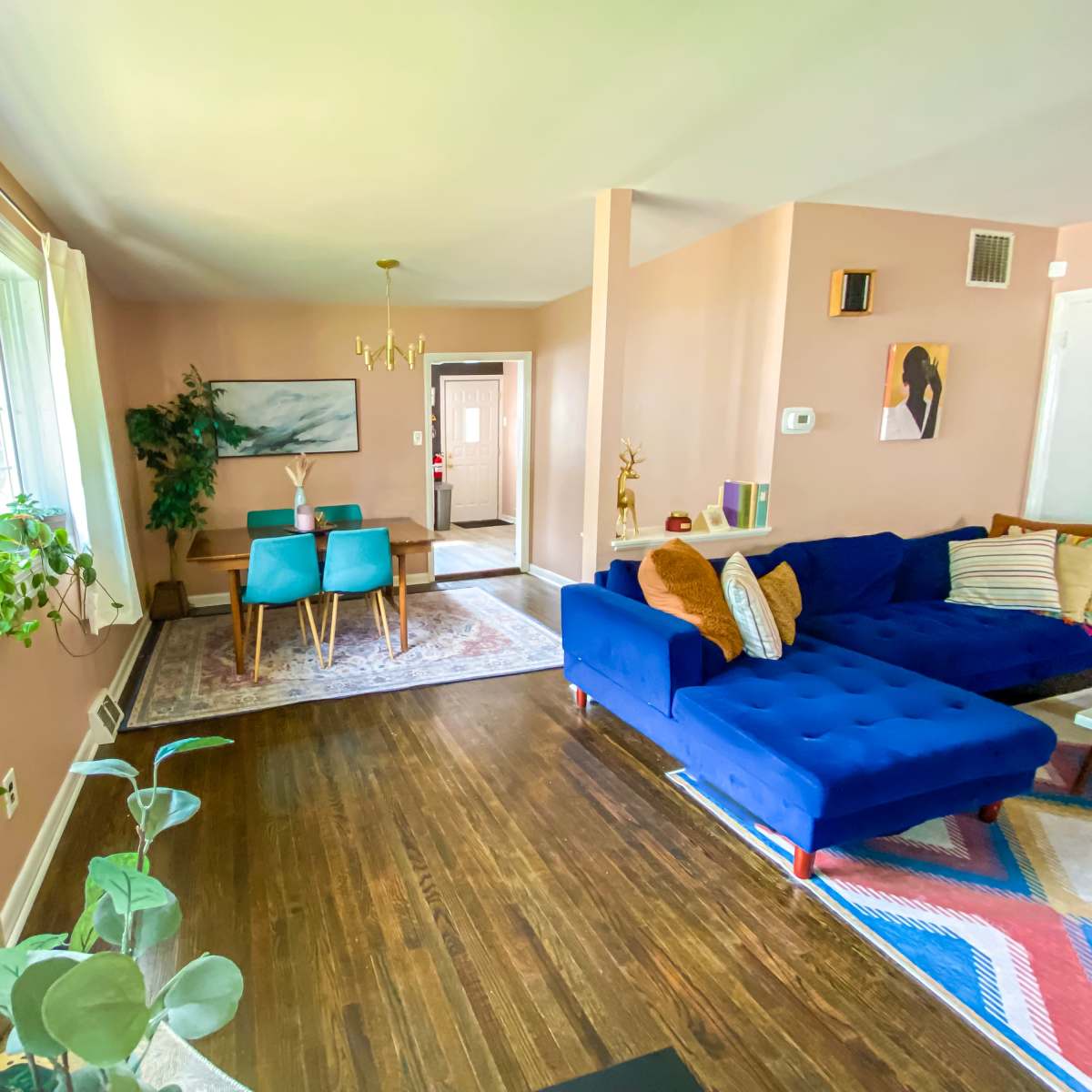 A cozy living room features a blue sofa, a small wooden table with turquoise chairs, and large windows allowing natural light to enter.
