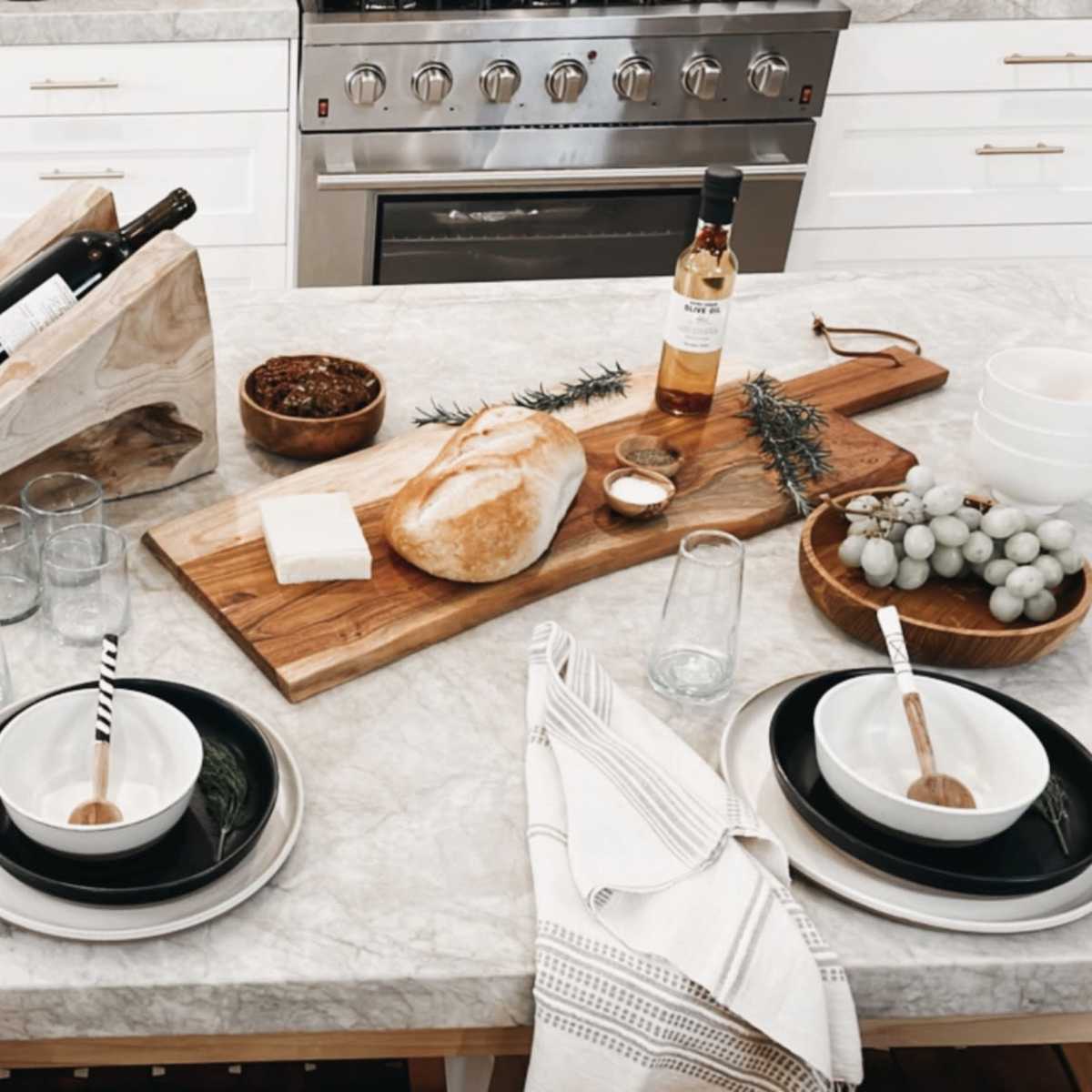 A kitchen table is set with black and white dishware, glasses, and a variety of food items, including bread, raspberries, and a bottle of wine, arranged on a marble countertop.