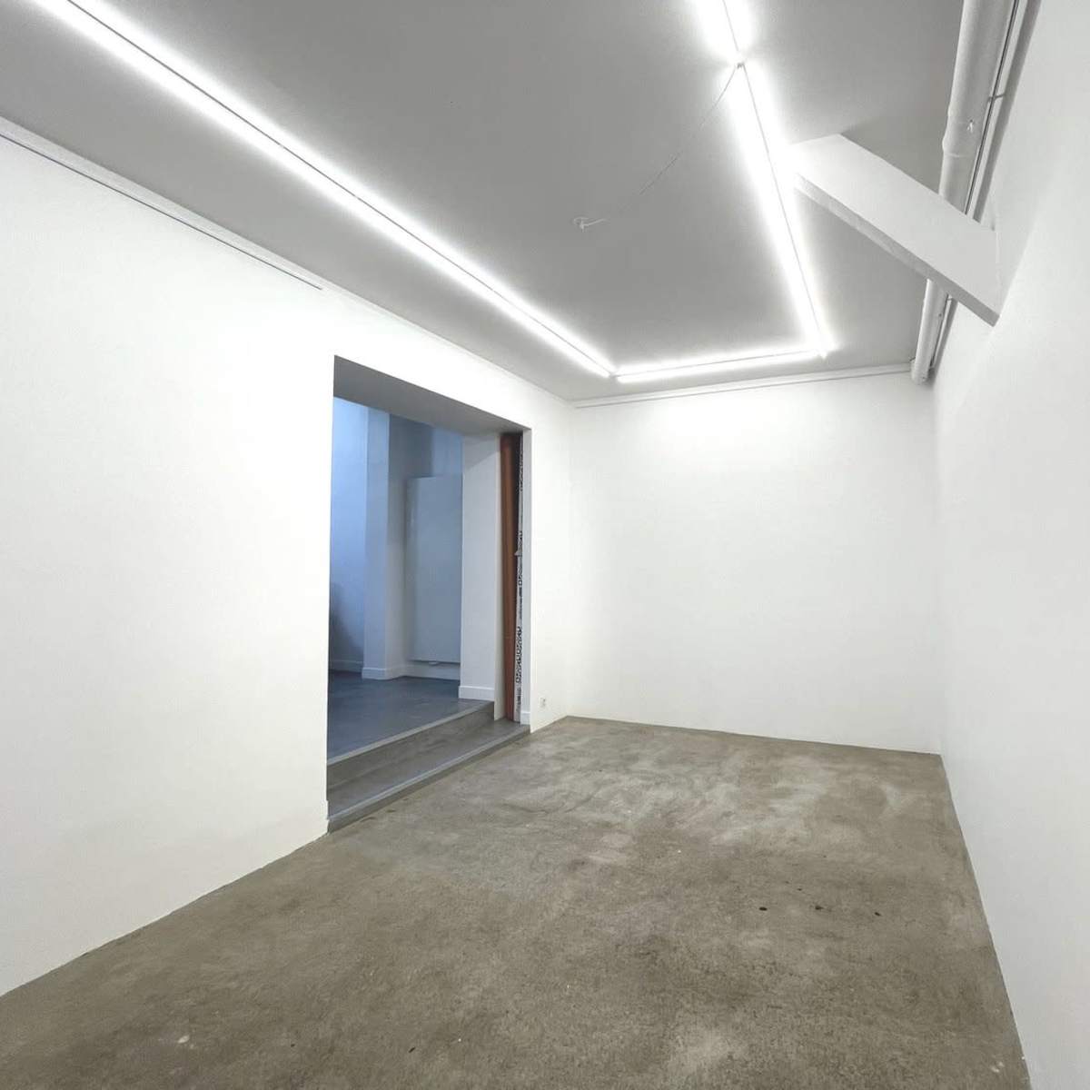 An empty room with a concrete floor and white walls features recessed lighting and an open doorway leading to a separate space.