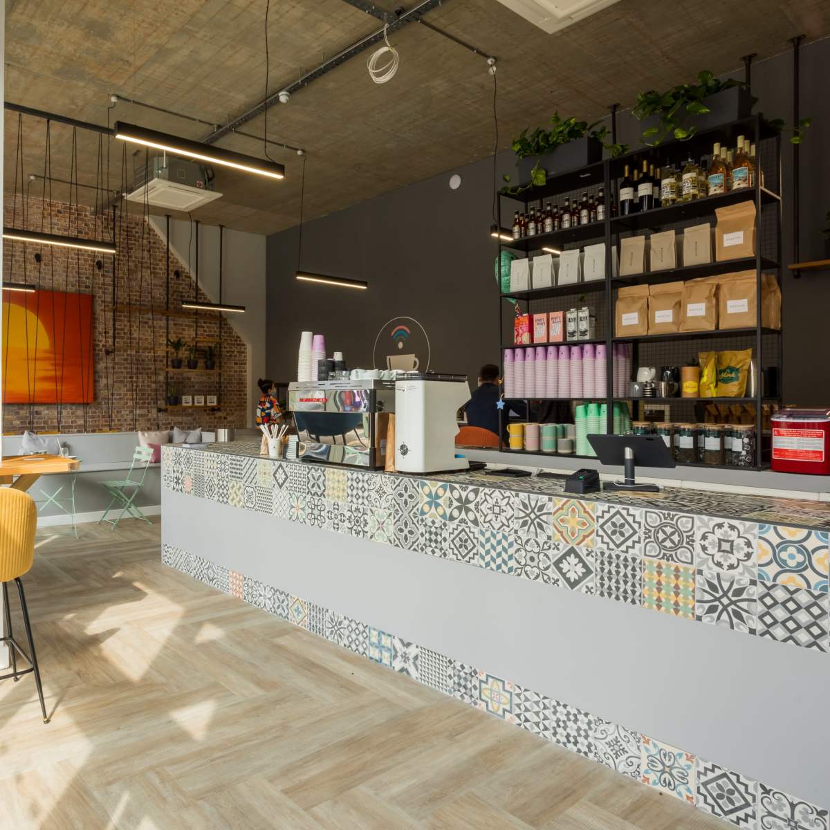A modern cafe interior with a patterned tile counter, wooden seating, and shelves filled with bottles and packaged goods.