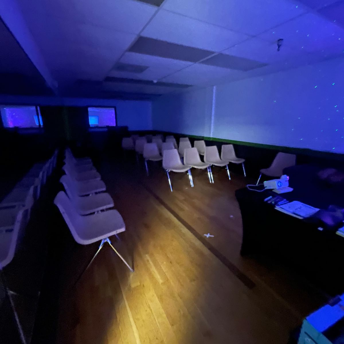 A dimly lit room with rows of empty chairs, a table in the foreground, and walls displaying a faint star-like projection.