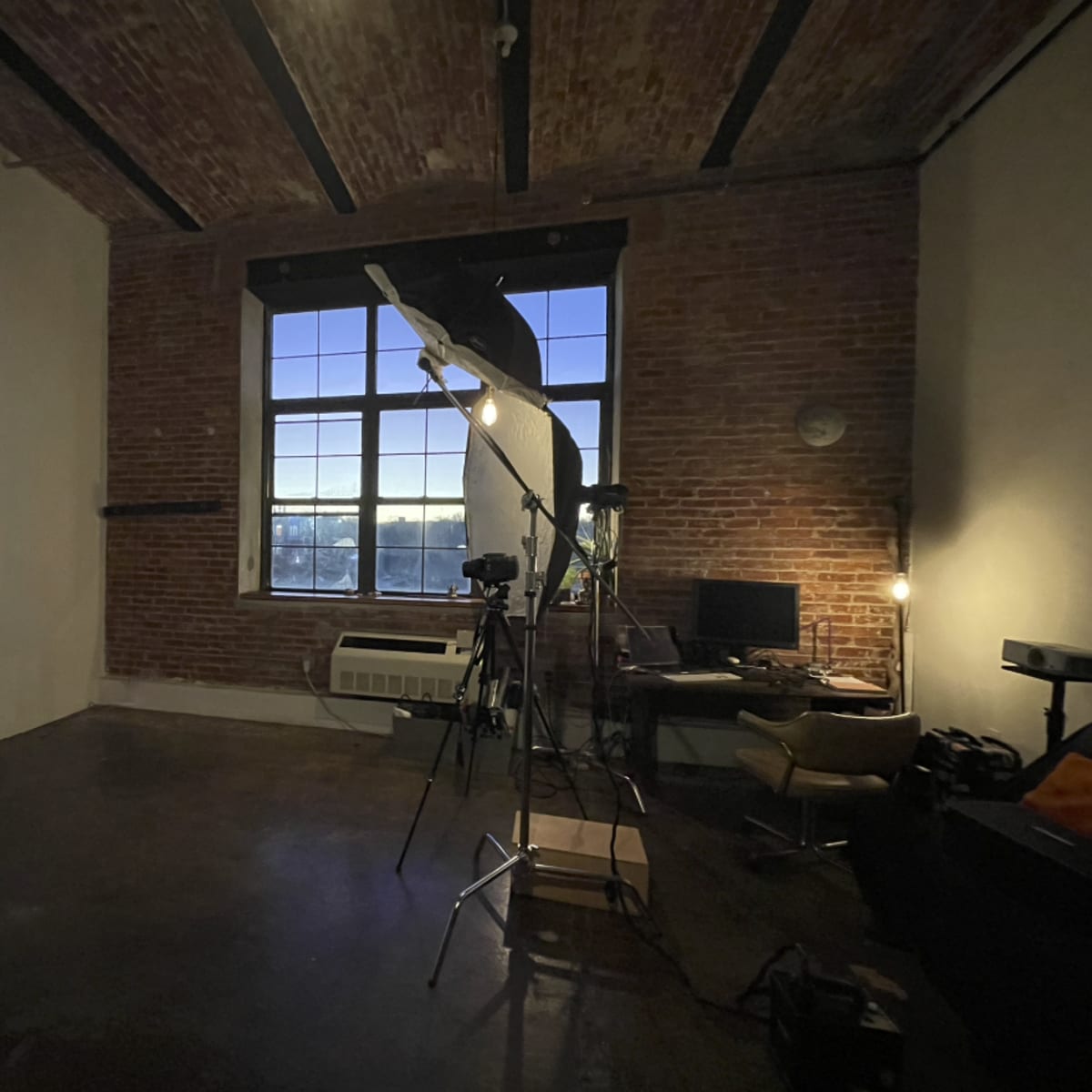 A spacious room with exposed brick walls, a large window, and photography equipment set up near a chair and a desk.