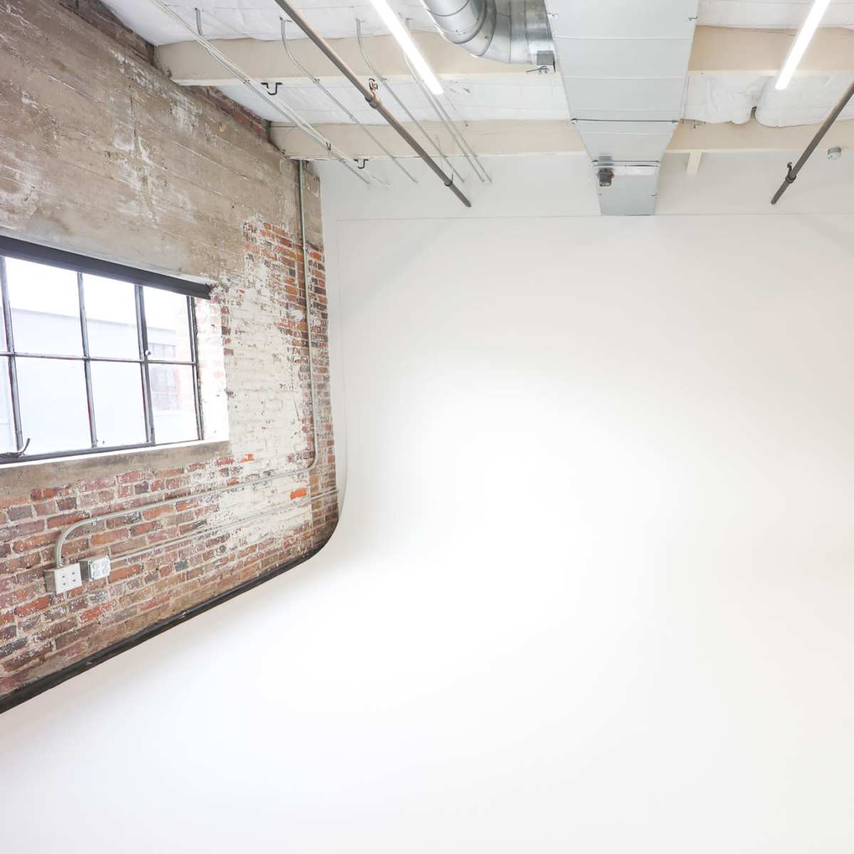 A spacious photography studio with a white backdrop, exposed brick walls, and a few pieces of furniture in the corner.