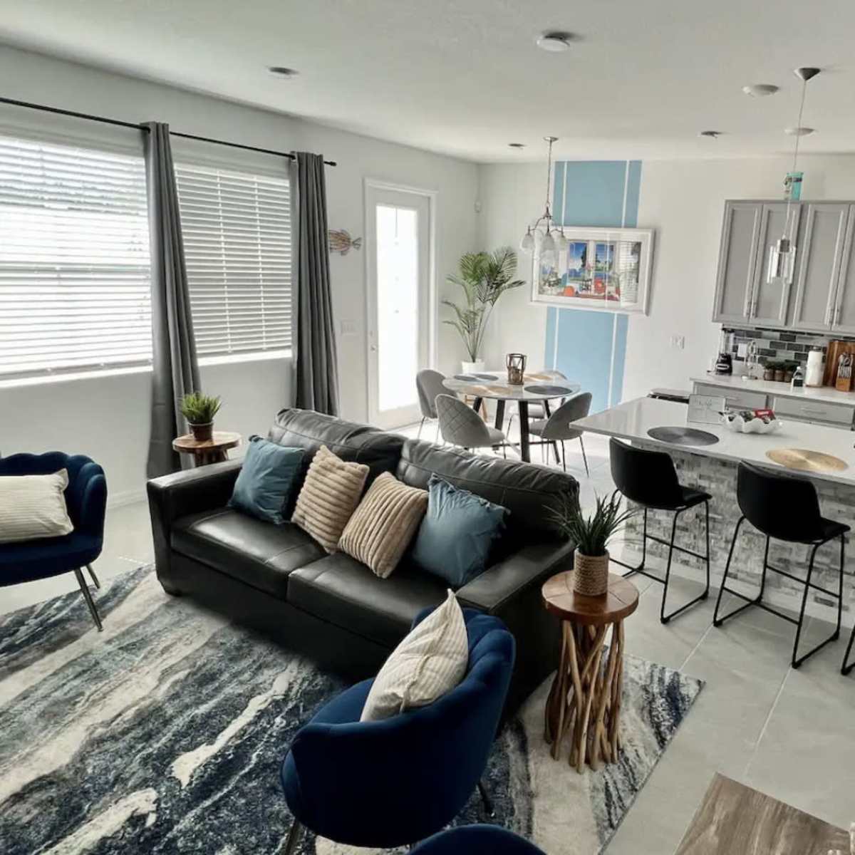 A modern living space featuring a gray sofa, blue chairs, a dining area, and a kitchen with stainless steel appliances.