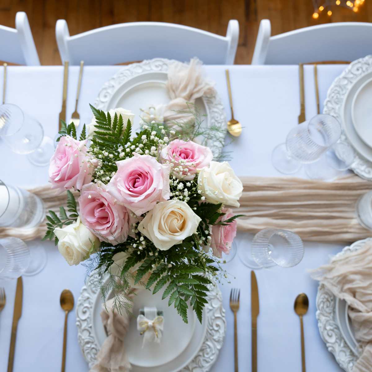 A beautifully set dining table features white plates with ornate edges, gold cutlery, and a centerpiece of pink and white roses among greenery.
