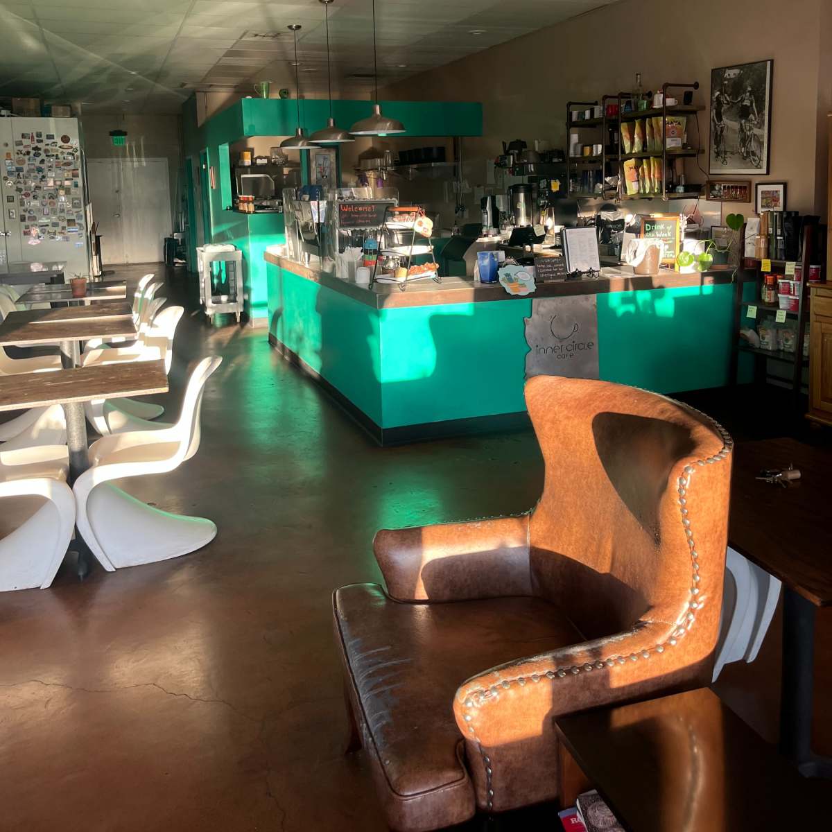A cozy coffee shop with a turquoise counter, wooden tables, and white chairs, illuminated by natural light.