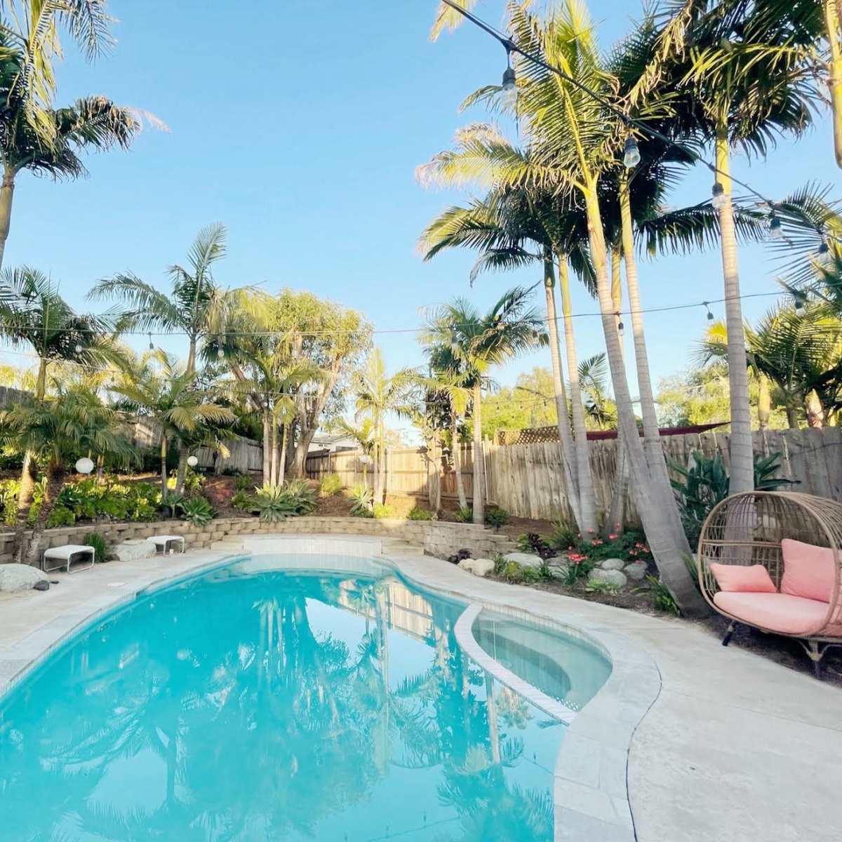 A swimming pool surrounded by palm trees and tropical plants, with two hanging chairs and a stone pathway nearby.