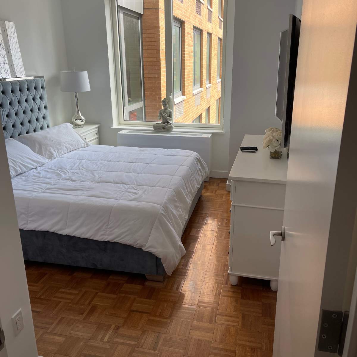 A neatly arranged bedroom features a bed with a gray headboard, a white comforter, a nightstand with a lamp, and a window showing a brick building outside.