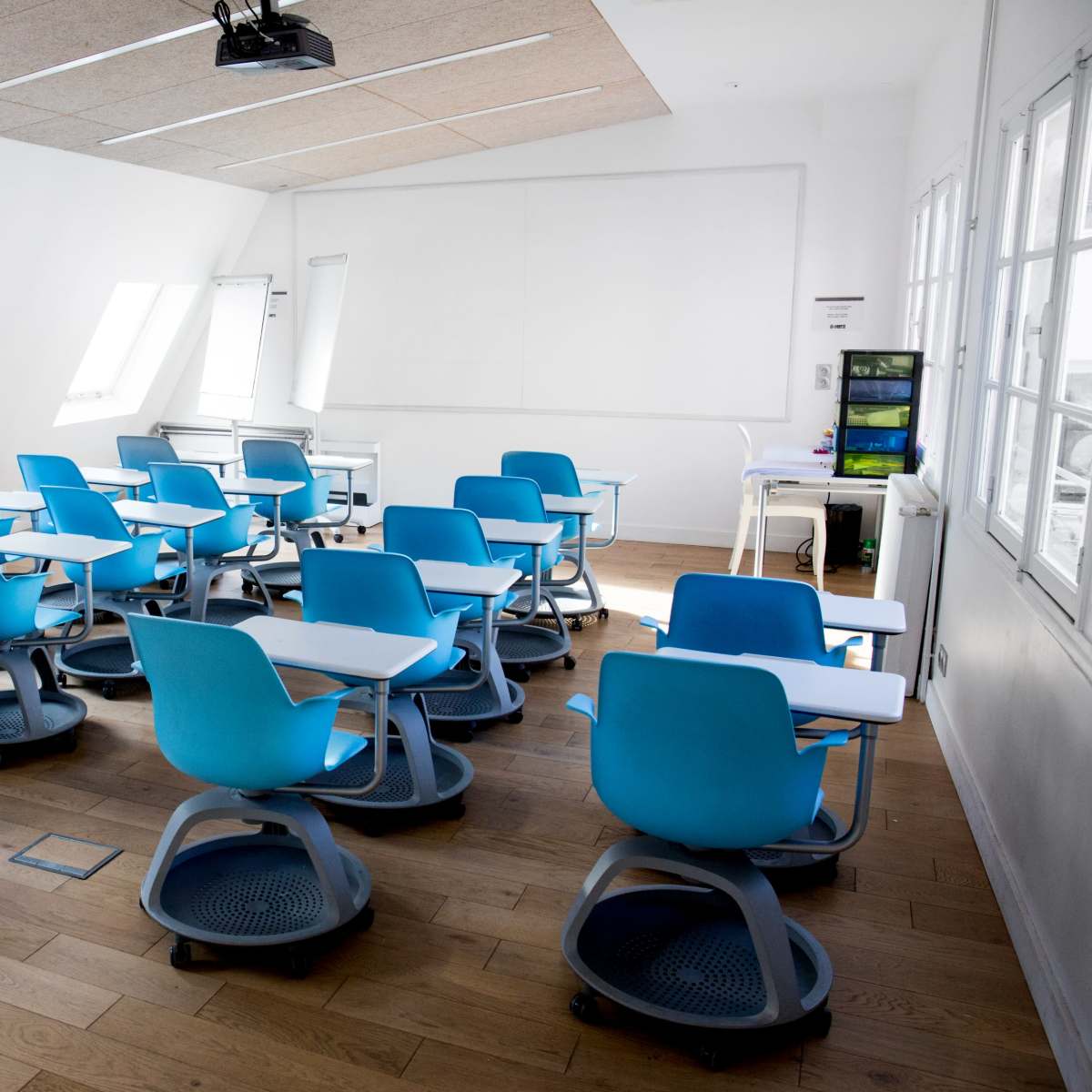A classroom is set up with blue rolling chairs arranged around small desks, and large windows allowing natural light to fill the space.