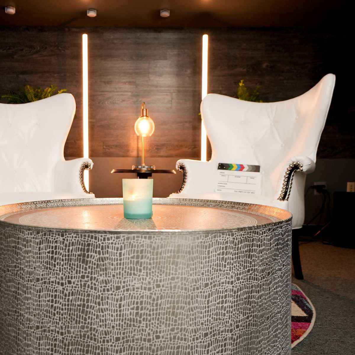 A living room setup featuring two white chairs, a decorative table with a candle, and illuminated wall fixtures in the background.