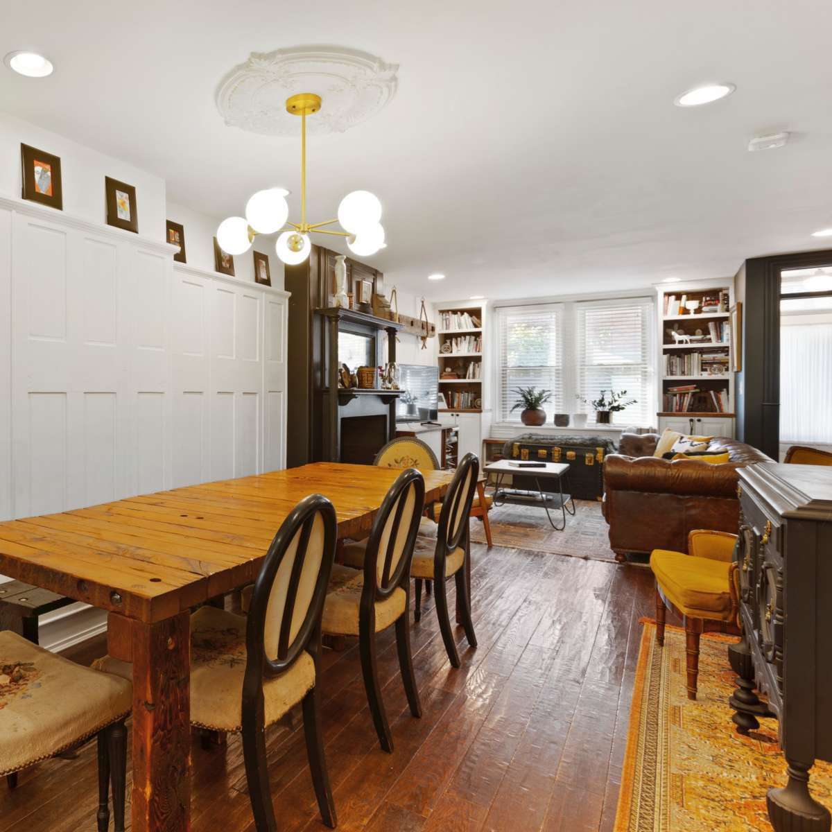 A dining area with a wooden table surrounded by six chairs, a large clock on the wall, and shelves filled with books in a well-lit room.