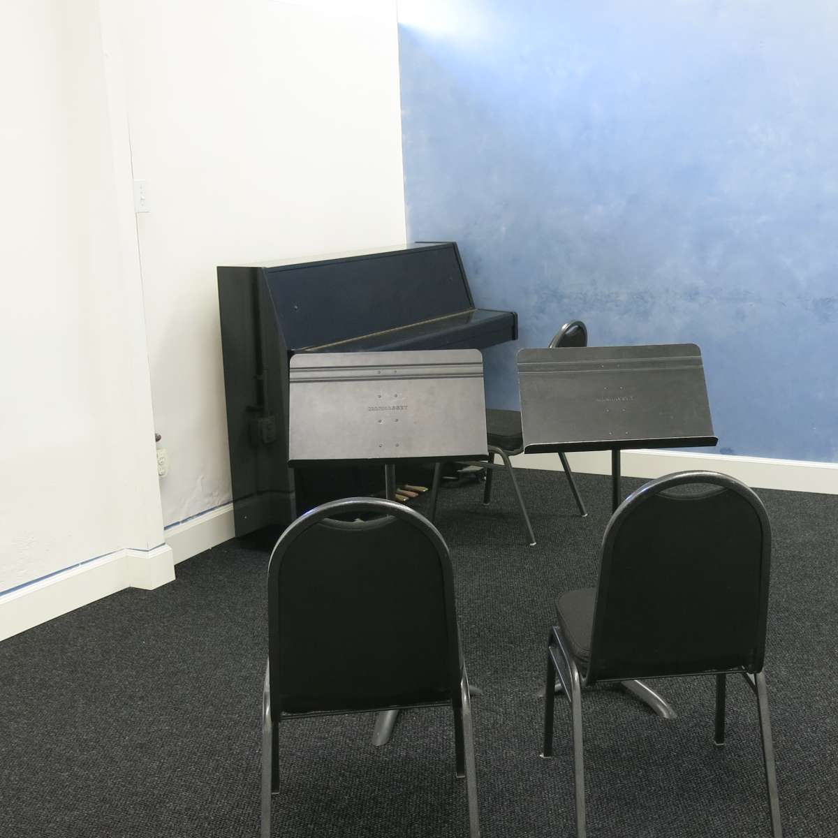A small room with two black chairs facing a blue wall, a stacked piano, and music stands in the corner.