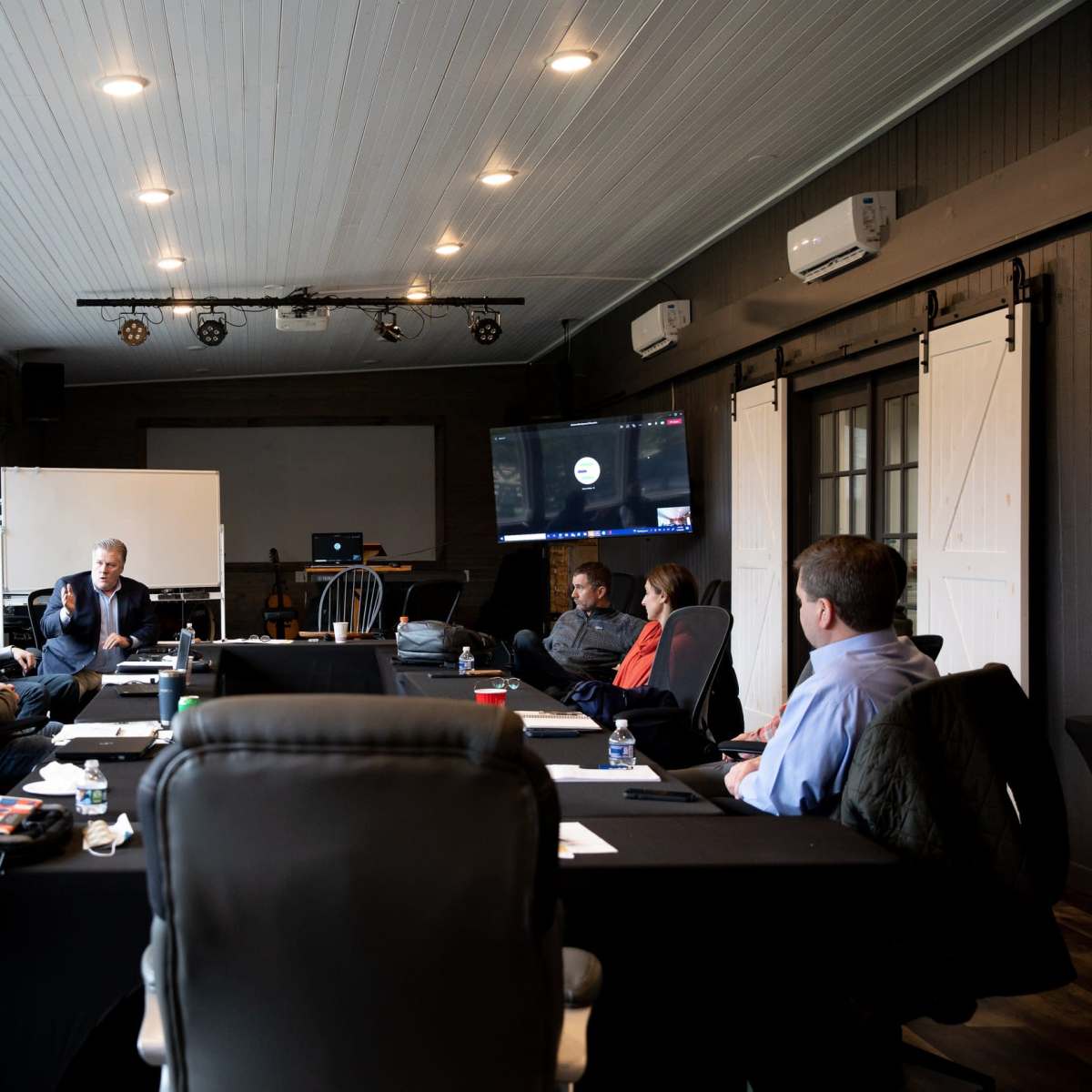 A group of eight people are seated around a black table in a conference room, engaging in a discussion, with a projection screen and whiteboard visible in the background.