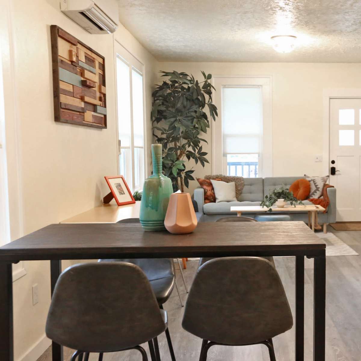 A cozy living area features a dining table with chairs, a blue sofa, a wooden shelf, and a plant near a front door with a window.