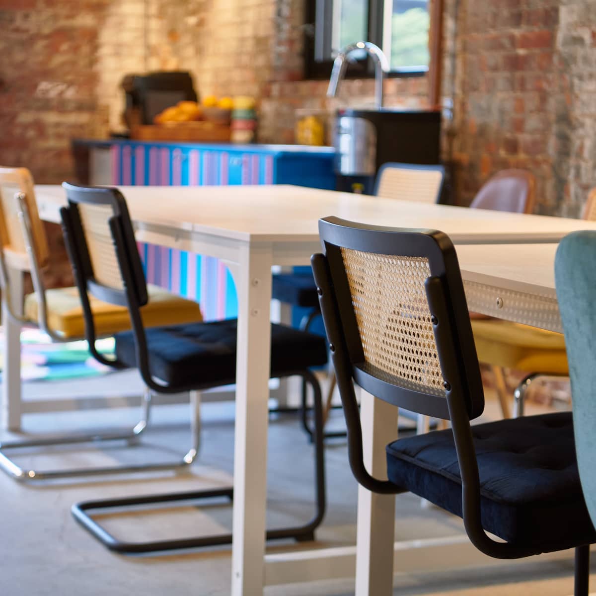 A modern workspace features a white table surrounded by various chairs in front of a brick wall and a colorful counter with kitchen items.