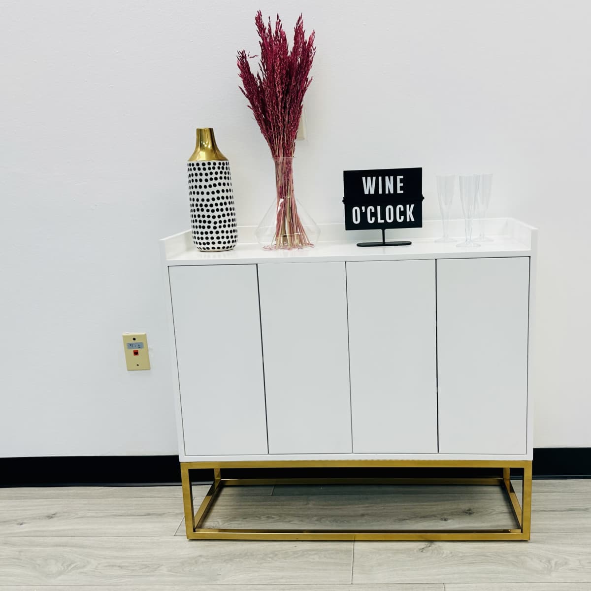 A white cabinet with gold accents holds a sign that reads "WINE O'CLOCK," accompanied by a vase and two glasses.