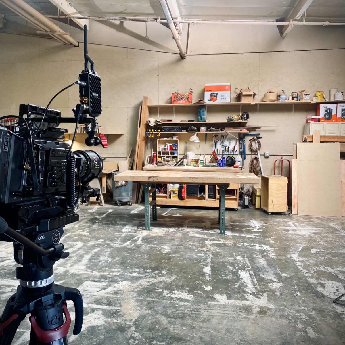 A video camera is positioned in a workshop facing a large wooden table, with shelves filled with tools and materials in the background.