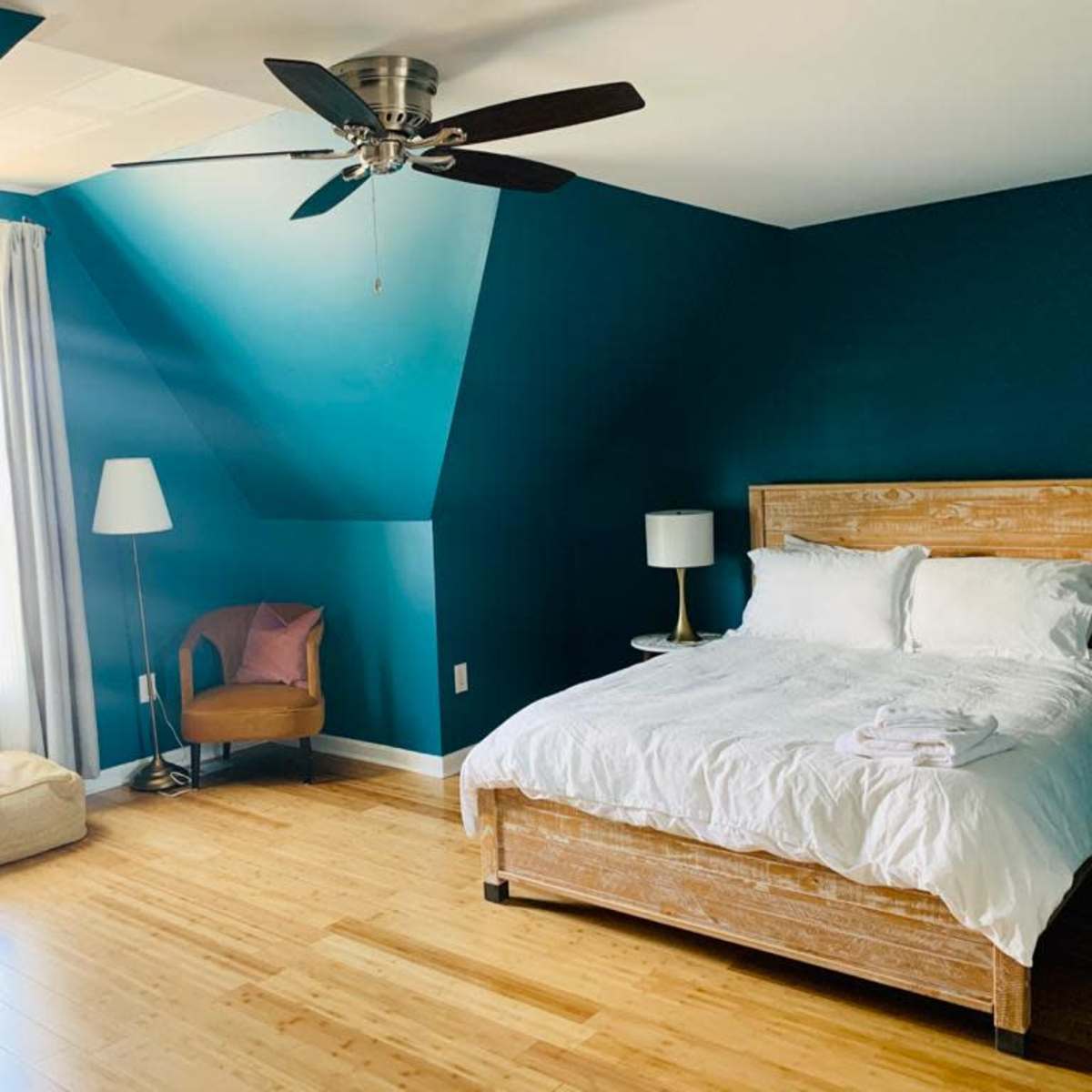 A bright bedroom features teal walls, a wooden bed with white linens, a chair, a lamp, and bamboo flooring.