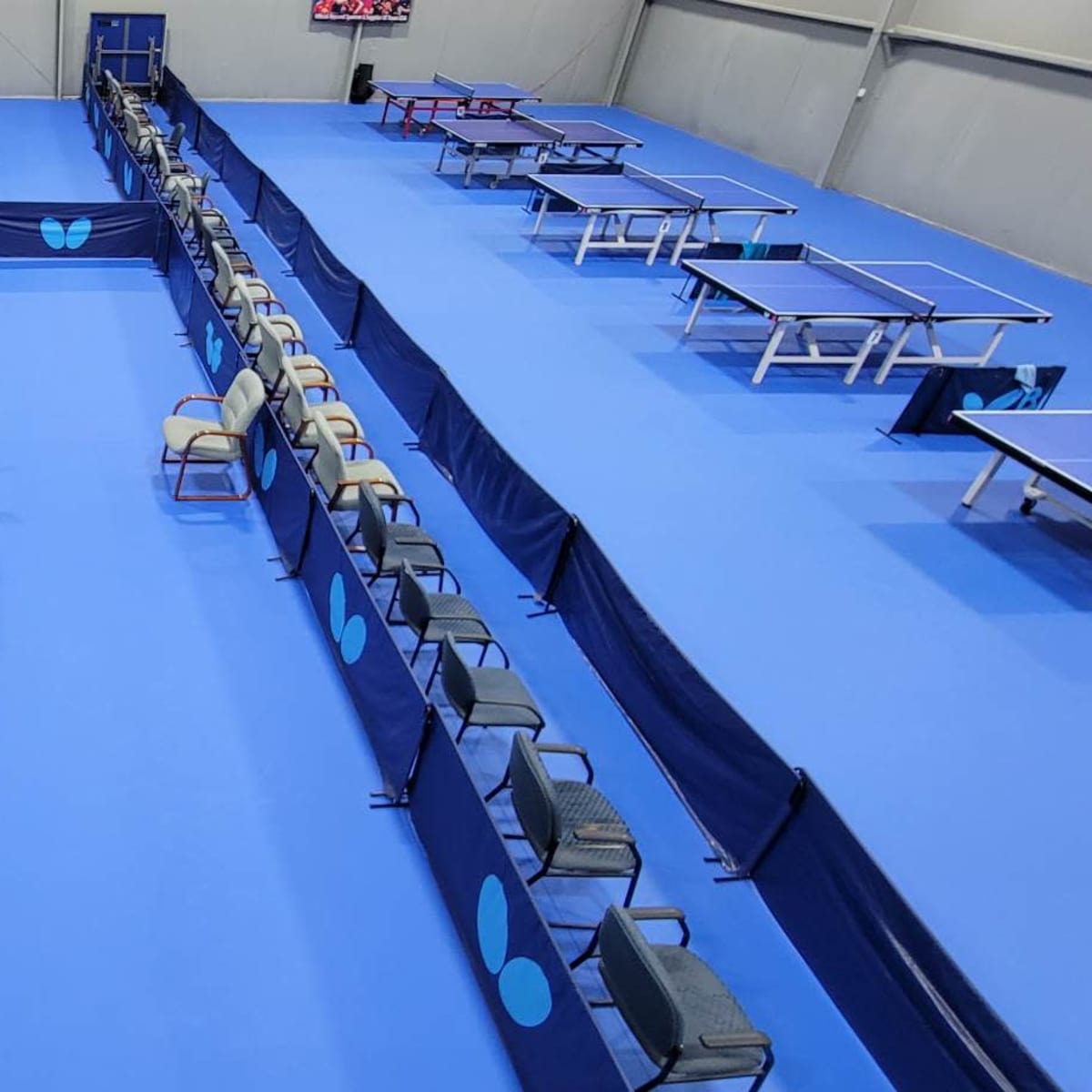 A spacious indoor table tennis facility with multiple blue tables, chairs arranged along the sides, and a wall displaying trophies.