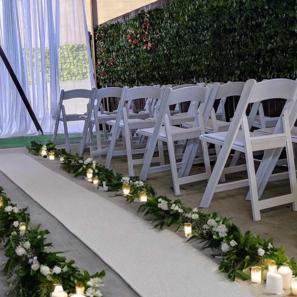 A wedding ceremony setup with white chairs arranged along a flower-lined aisle illuminated by candles.