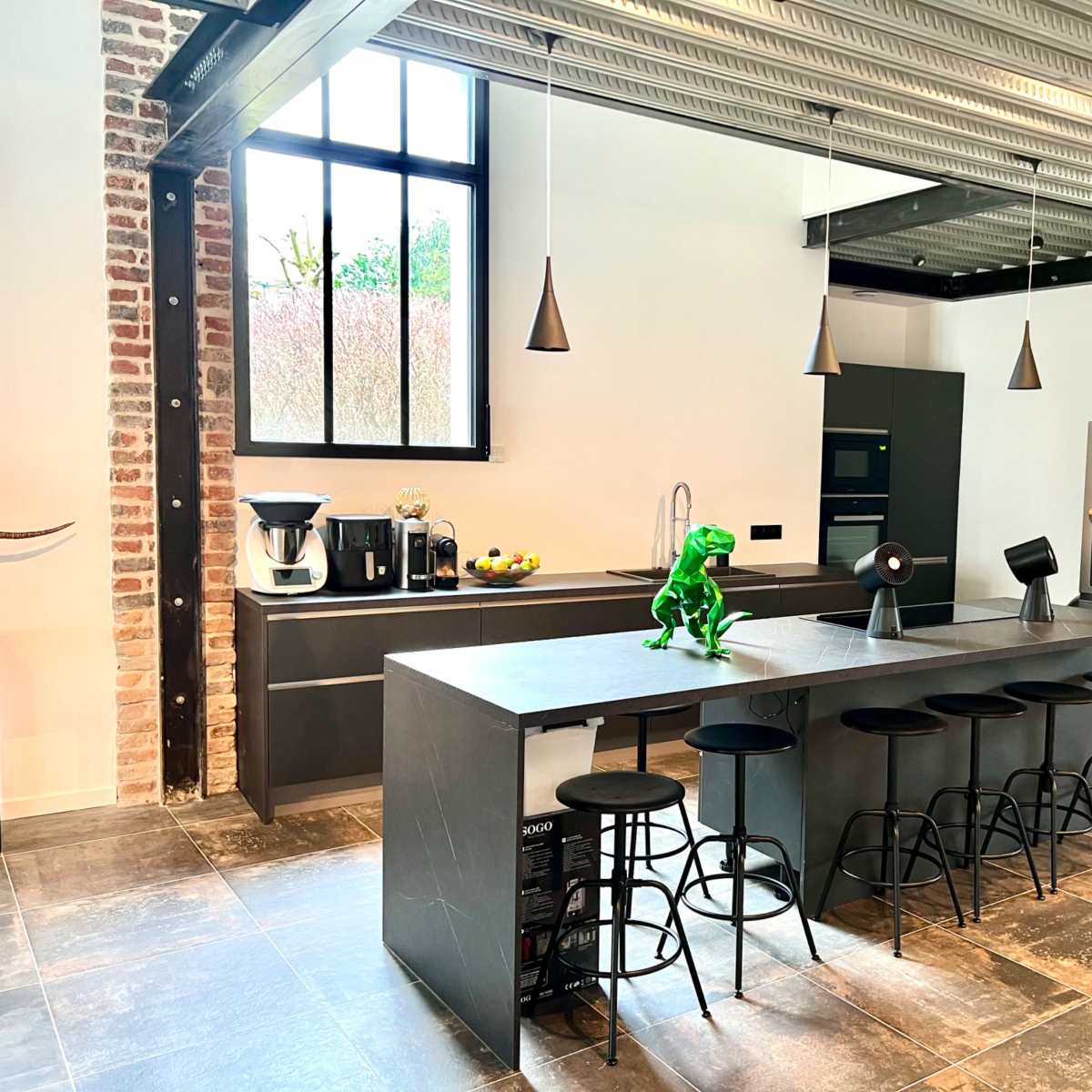 A modern kitchen with a long dark countertop, bar stools, and appliances including a coffee maker and refrigerator, all set against brick and metal accents.