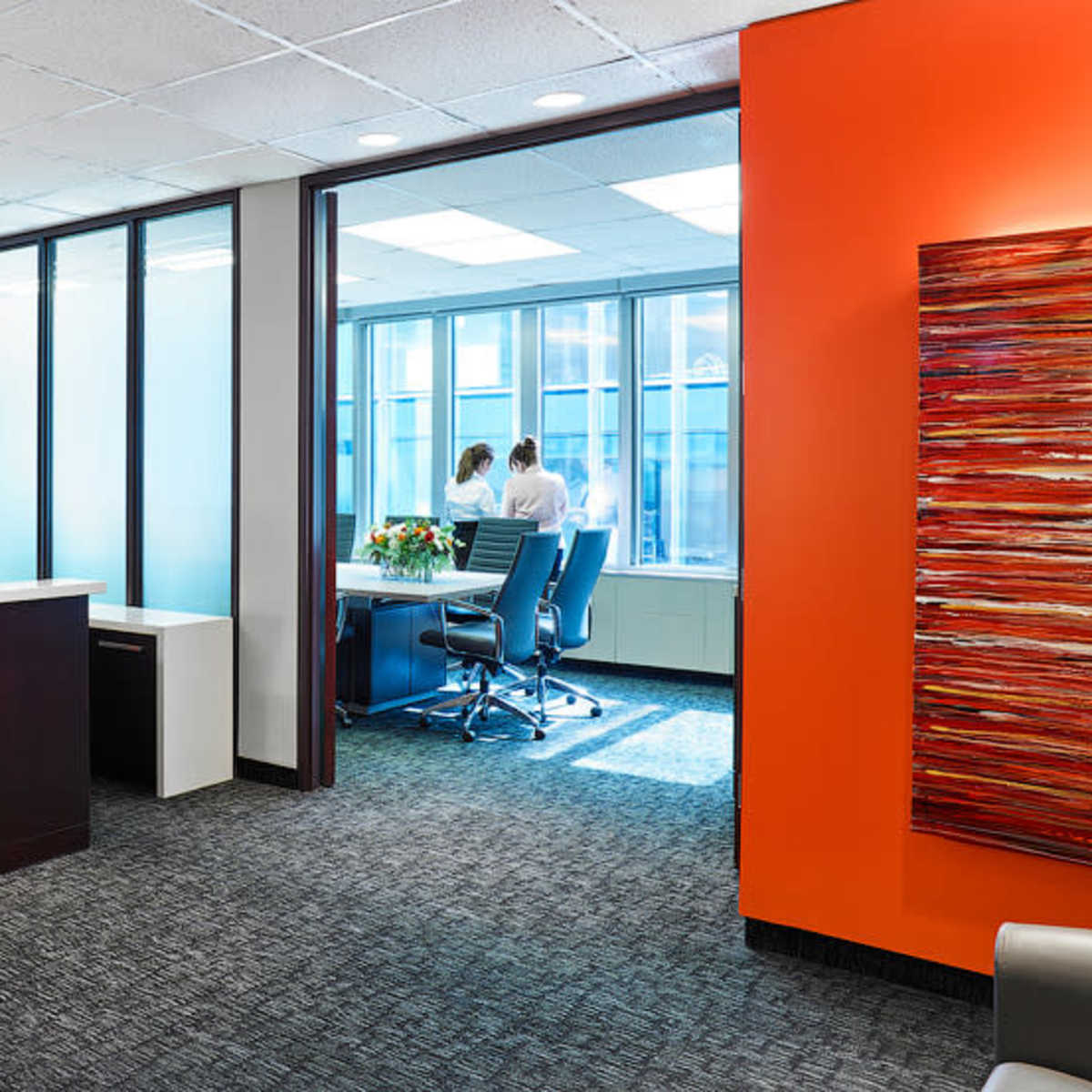 A modern office reception area with a front desk and a glass partition leading to a meeting room where two people are standing by a window.