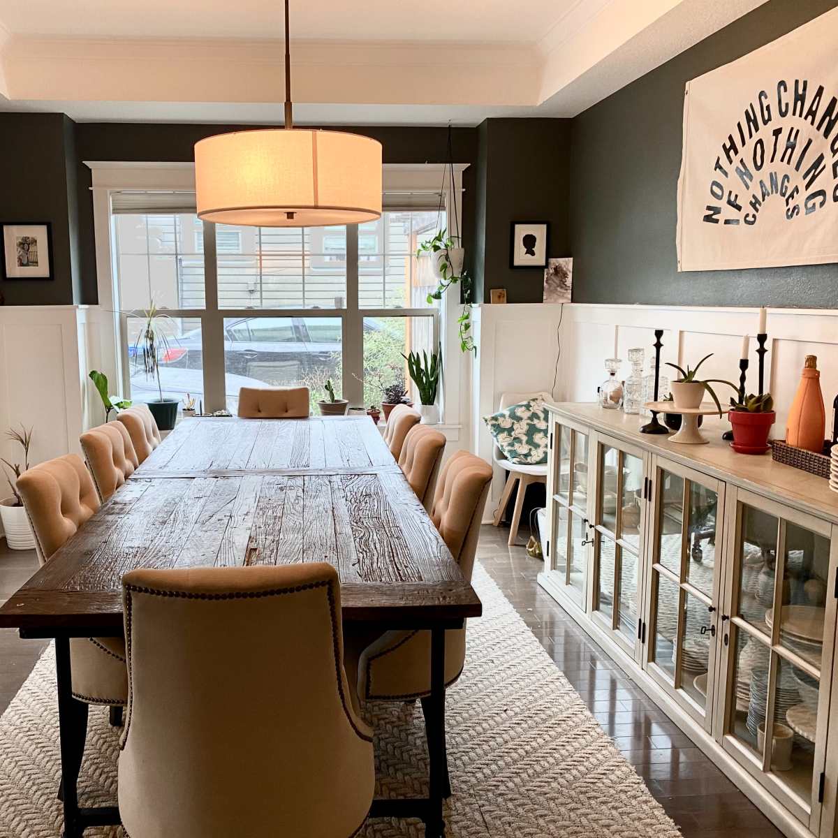 A spacious dining room features a long wooden table surrounded by upholstered chairs, with a large window providing natural light and shelves displaying various decorative items.