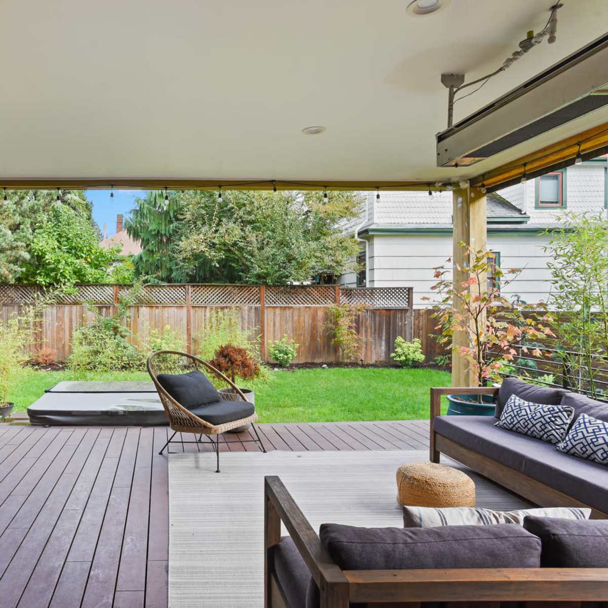 A modern outdoor patio with a wooden dining table, black chairs, a lounge area with a sofa and chair, and a well-maintained garden in the background.