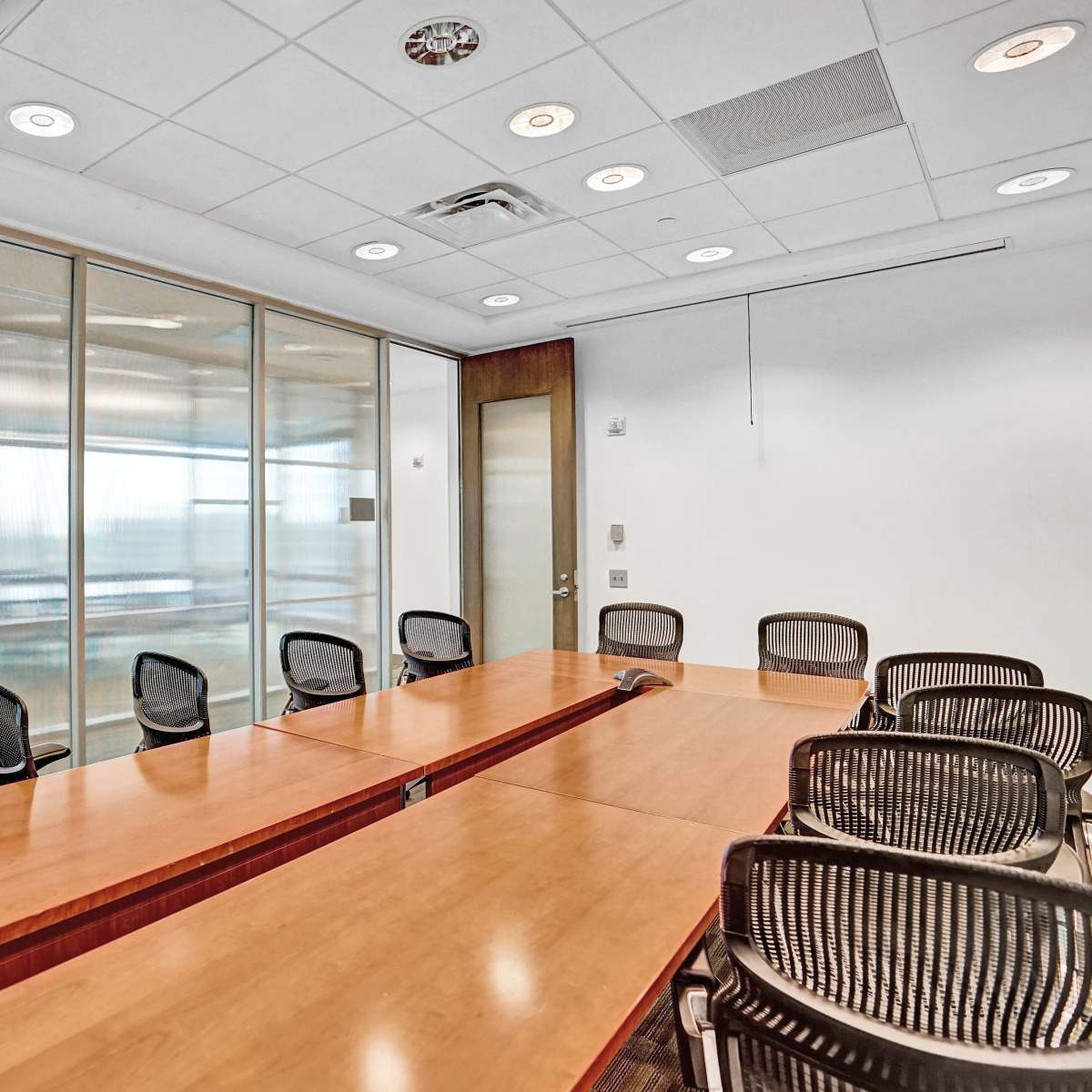 A conference room features a long wooden table surrounded by black mesh chairs and large windows that overlook an office view.