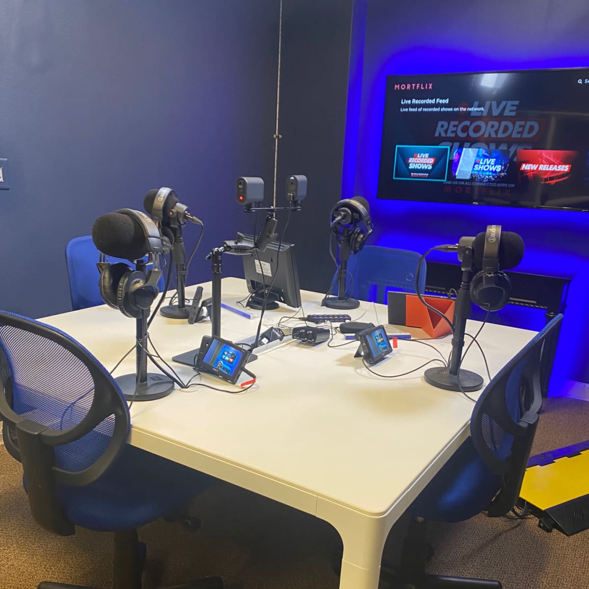 A recording studio with a round table surrounded by four chairs, each equipped with a microphone and headphones, alongside a monitor displaying the words "LIVE RECORDED."