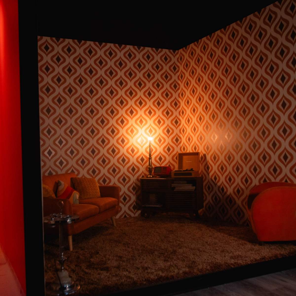 A vintage-inspired living room with geometric wall patterns, featuring a brown sofa, an orange armchair, a coffee table, and a retro-style television on a wooden console.