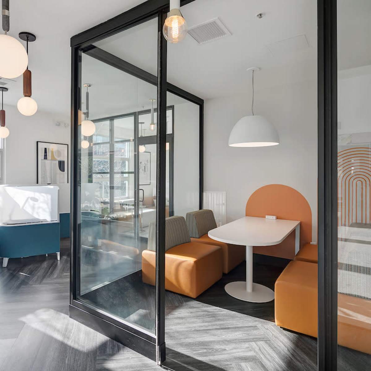 A modern meeting room with glass walls, a round table, orange seating, and pendant lights.
