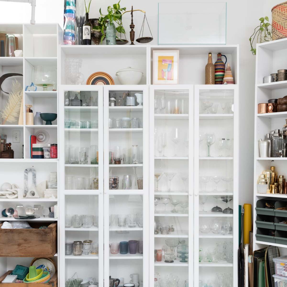 A well-organized display of glassware and decorative items in three white cabinets against a light-colored wall.