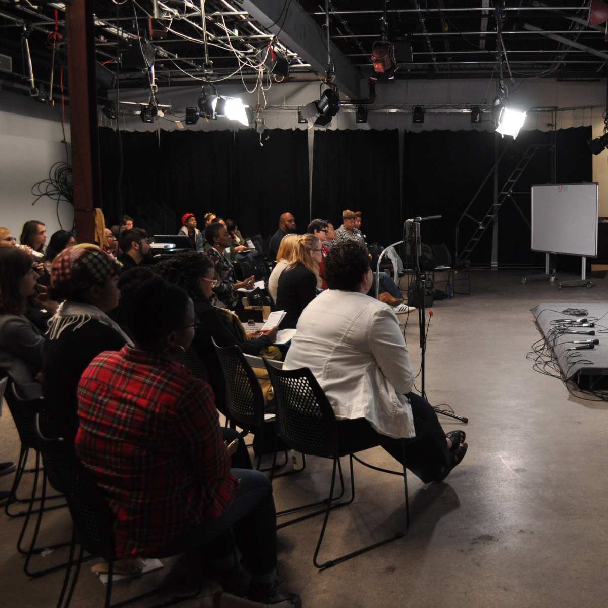 A panel discussion takes place on stage in a studio setting, with an audience seated in rows facing two speakers who are seated on stools.