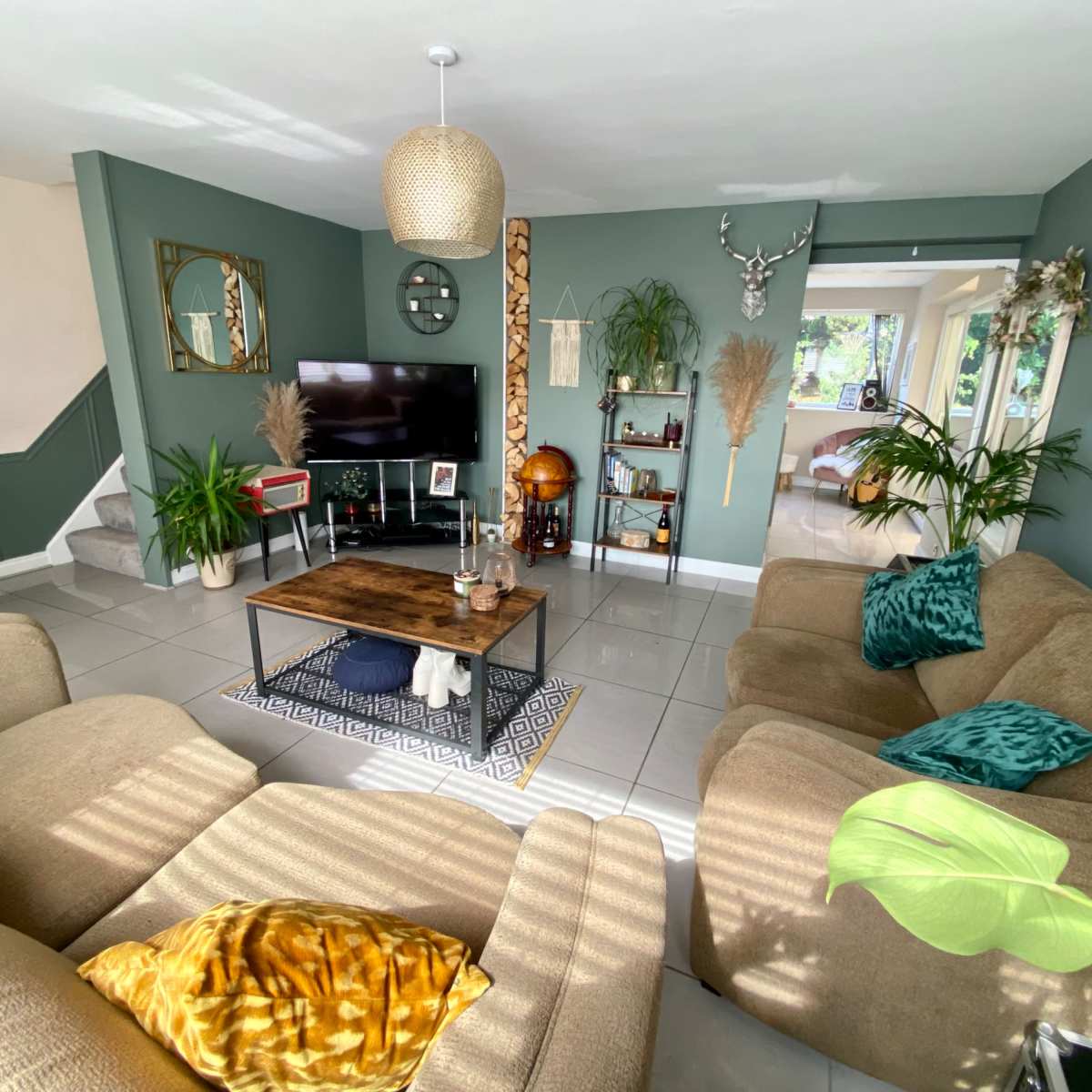 A cozy living room with beige sofas, a wooden coffee table, and various decorative plants, featuring a television on a wall surrounded by artwork.