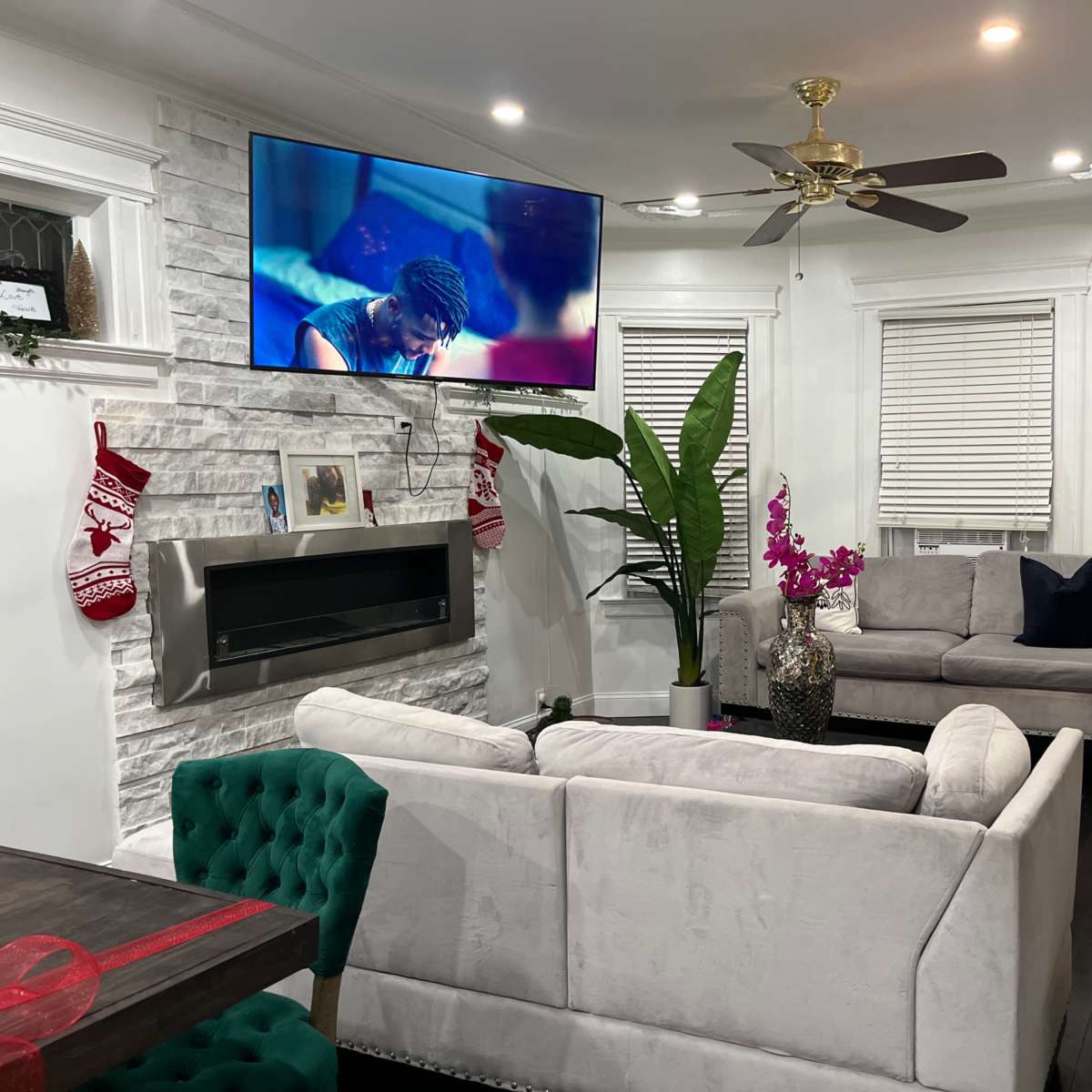 A living room features a television mounted on the wall, a modern gray sofa, a green accent chair, a decorative plant, and a fireplace with stockings hung nearby.