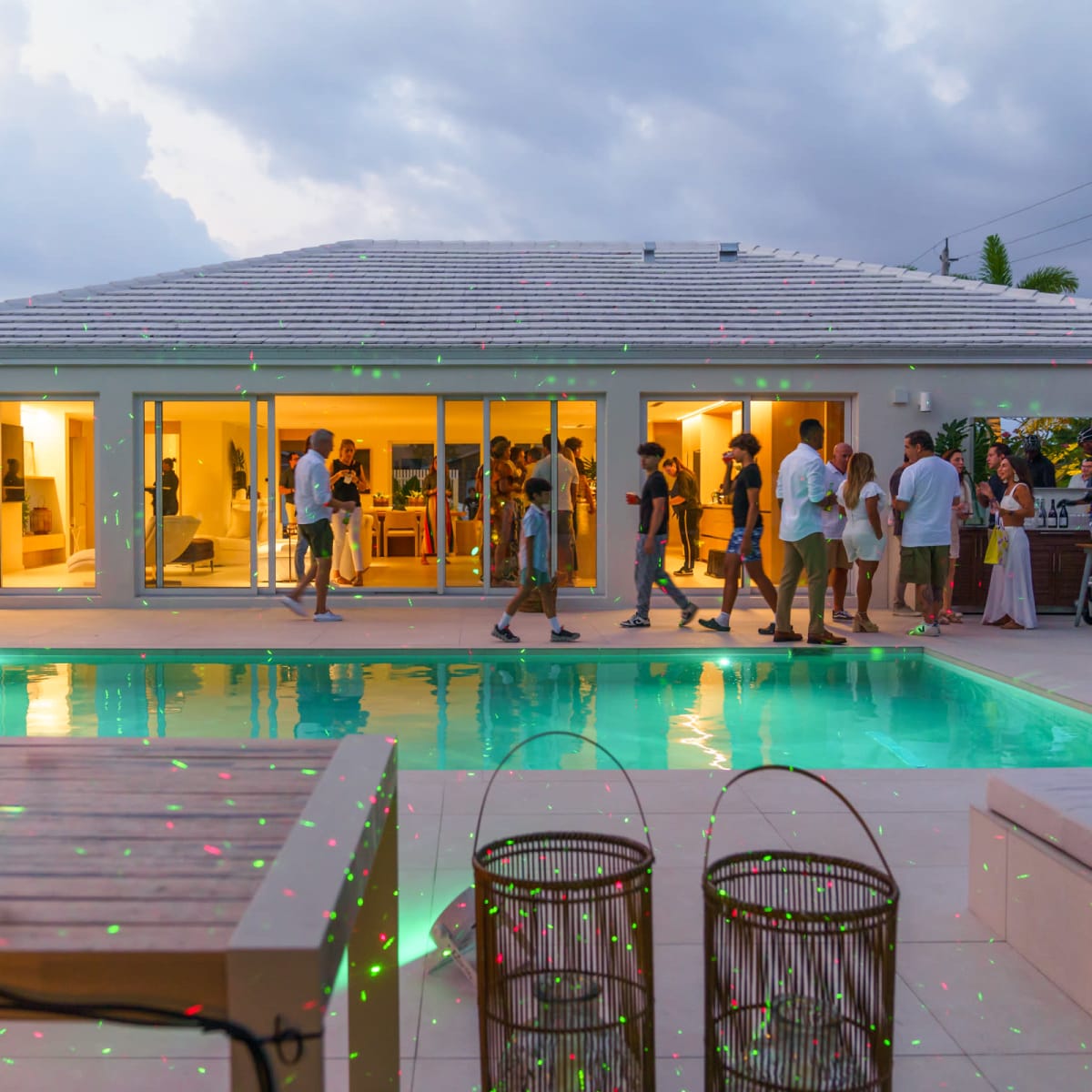 A group of people socializes around a pool at a modern house during an evening gathering.