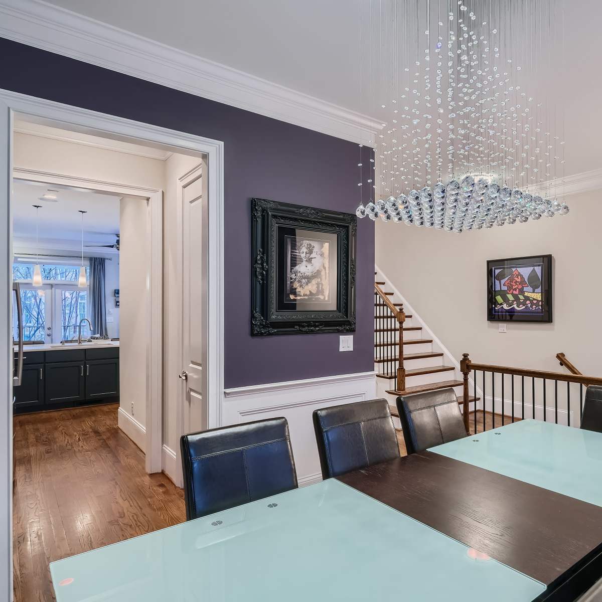 A modern dining room features a long glass table surrounded by black chairs, with a chandelier above and artwork on the walls.