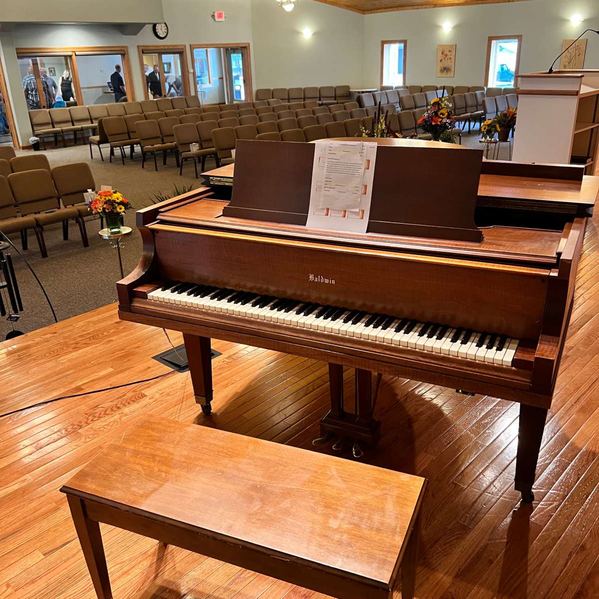 A wooden piano with a sheet of music sits on a stage in a spacious room filled with rows of chairs.