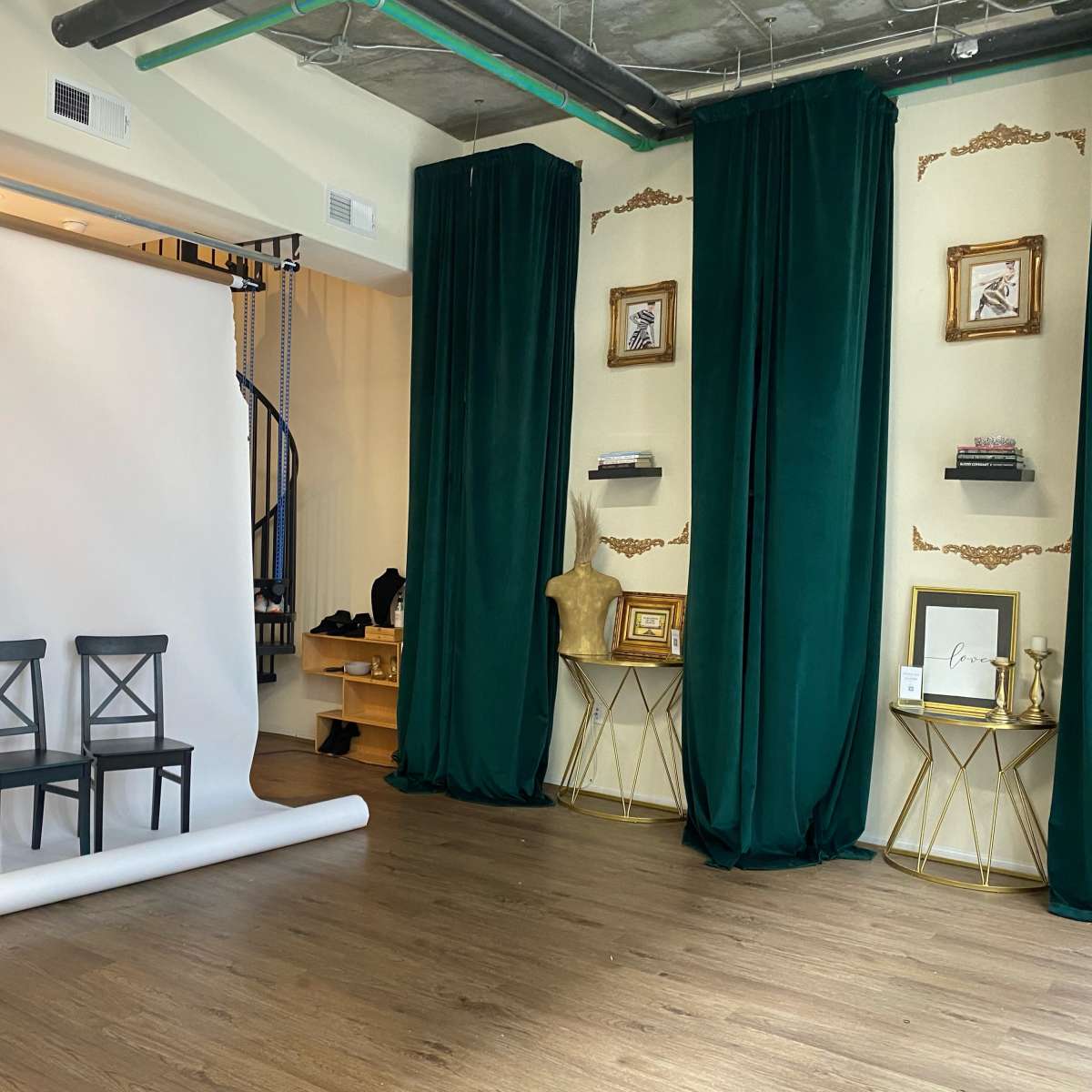 A spacious studio with a white backdrop, three black chairs in front, and green curtains draping the walls, alongside decorative shelves with various items.