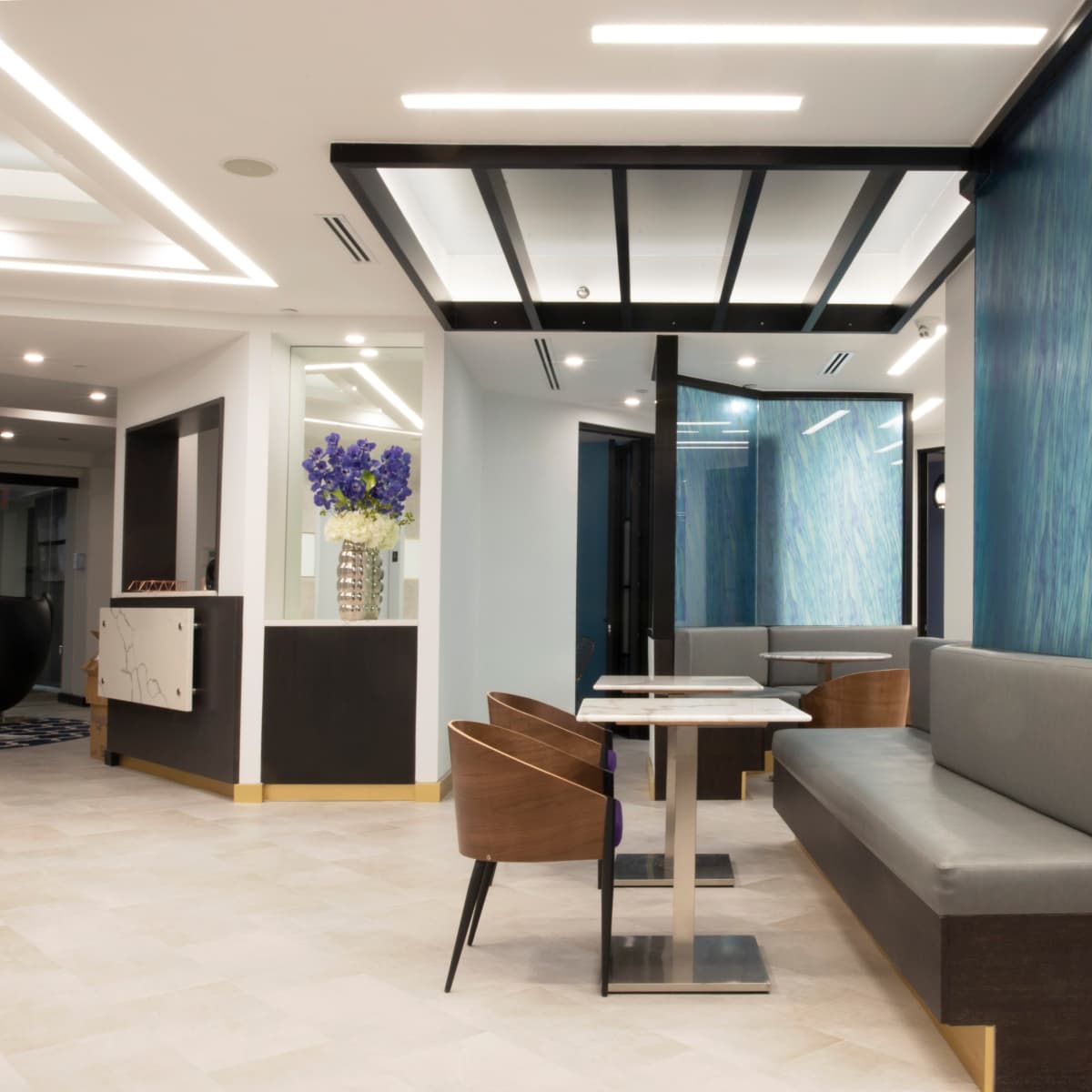 A modern interior space featuring a seating area with gray benches and wooden chairs, complemented by blue accent walls and decorative floral arrangements.