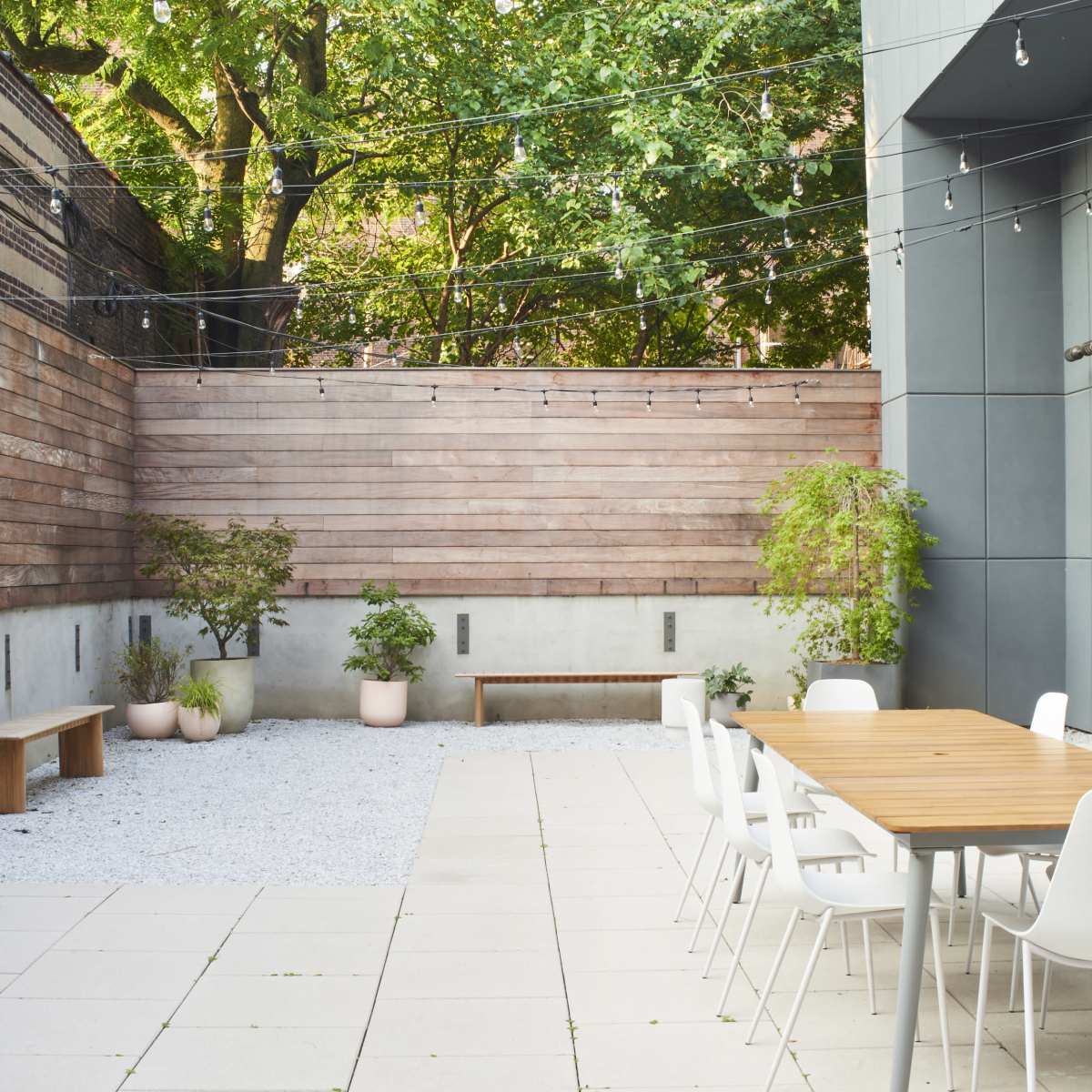 A modern outdoor patio area with a wooden table surrounded by white chairs, gravel ground, and green plants against a wood-paneled wall.