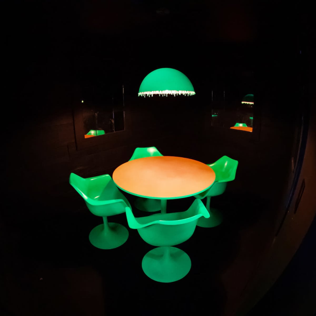 A round table with a light orange surface is surrounded by four green chairs beneath a green lamp in a dark room.