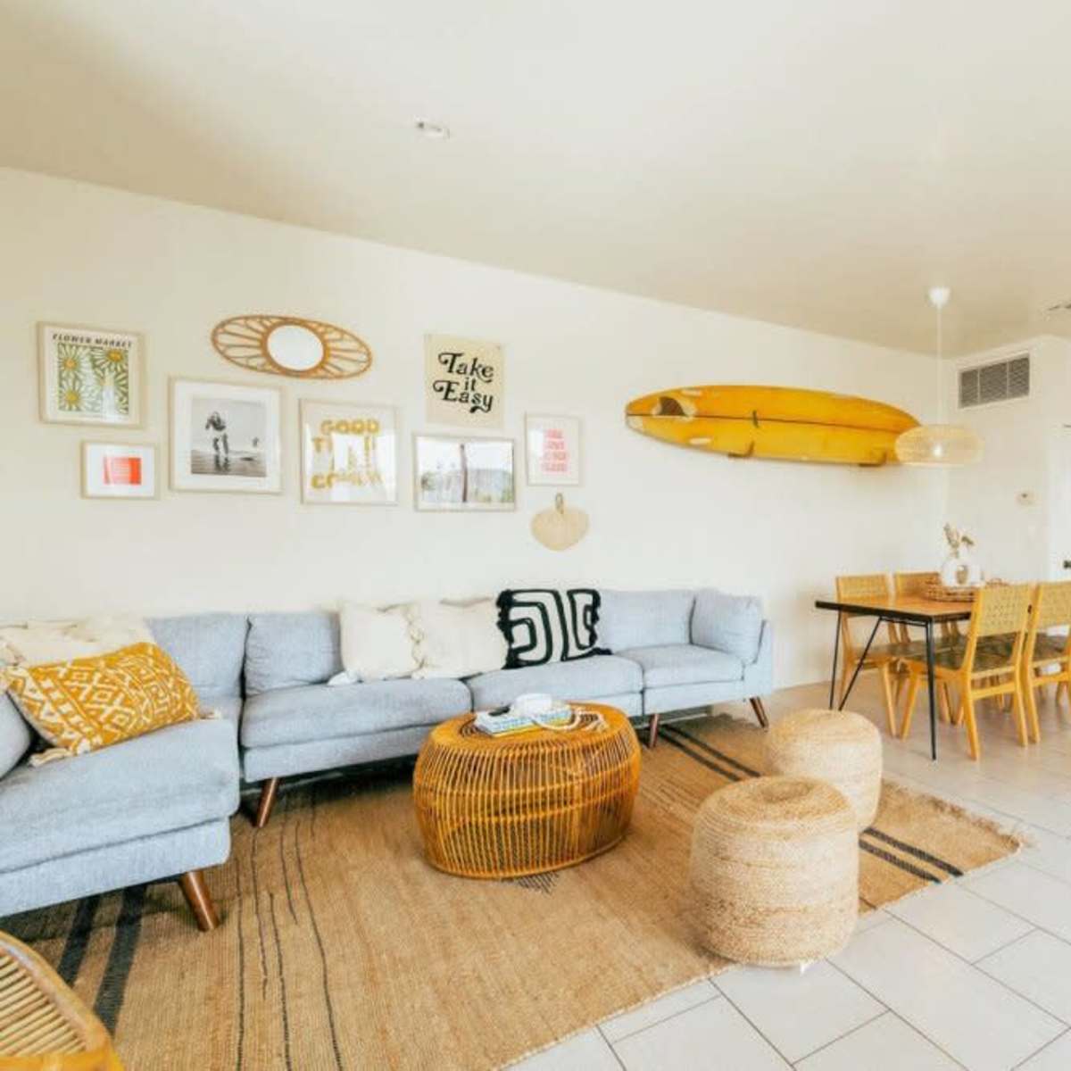 A modern living space featuring a light-colored sofa, a round rattan coffee table, dining chairs, a dining table, and a kitchen area with a surfboard mounted on the wall.